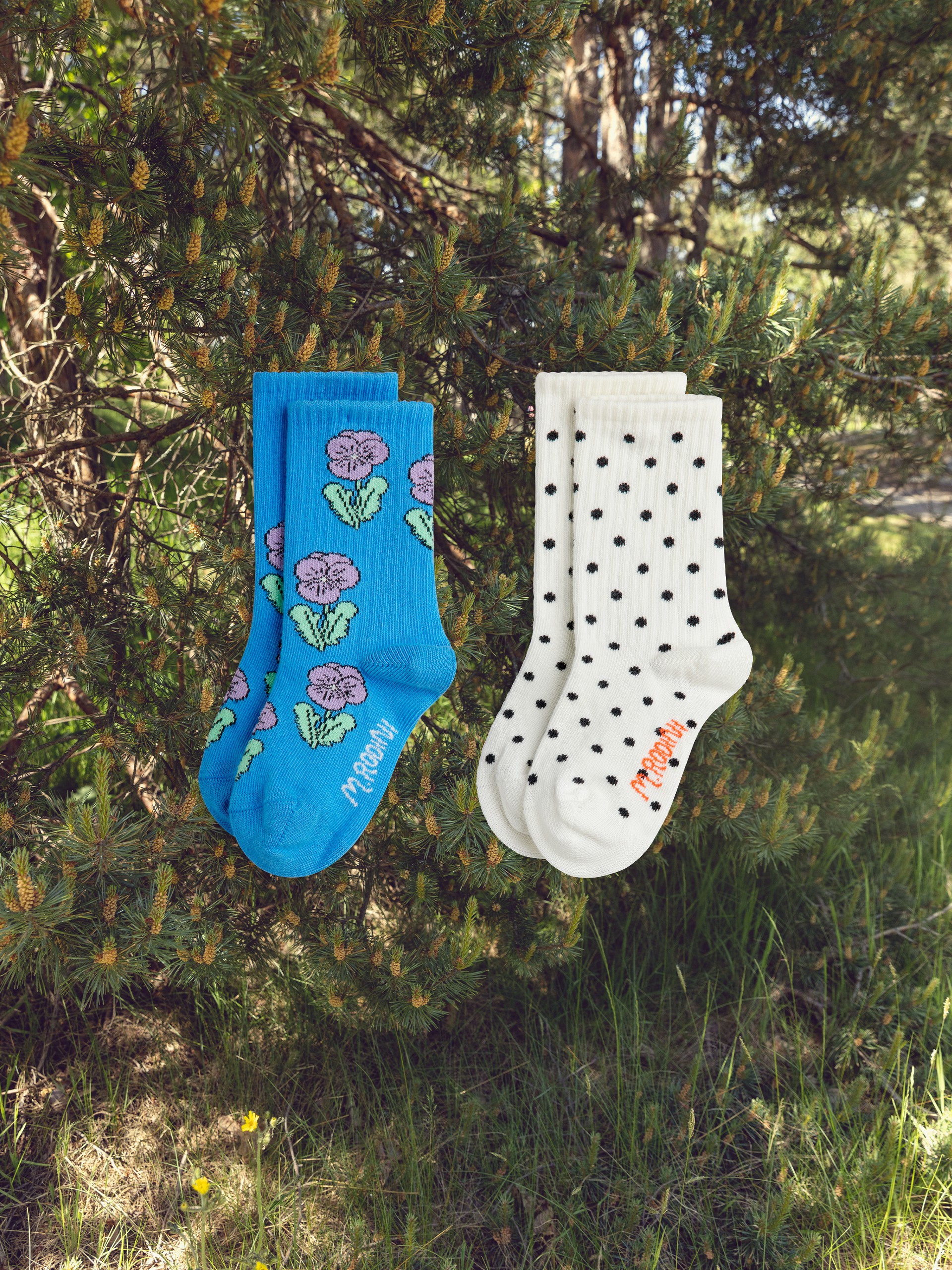 Two pairs of socks hang from tree branches: one blue with purple flowers, the other white with black polka dots, against a forest backdrop.