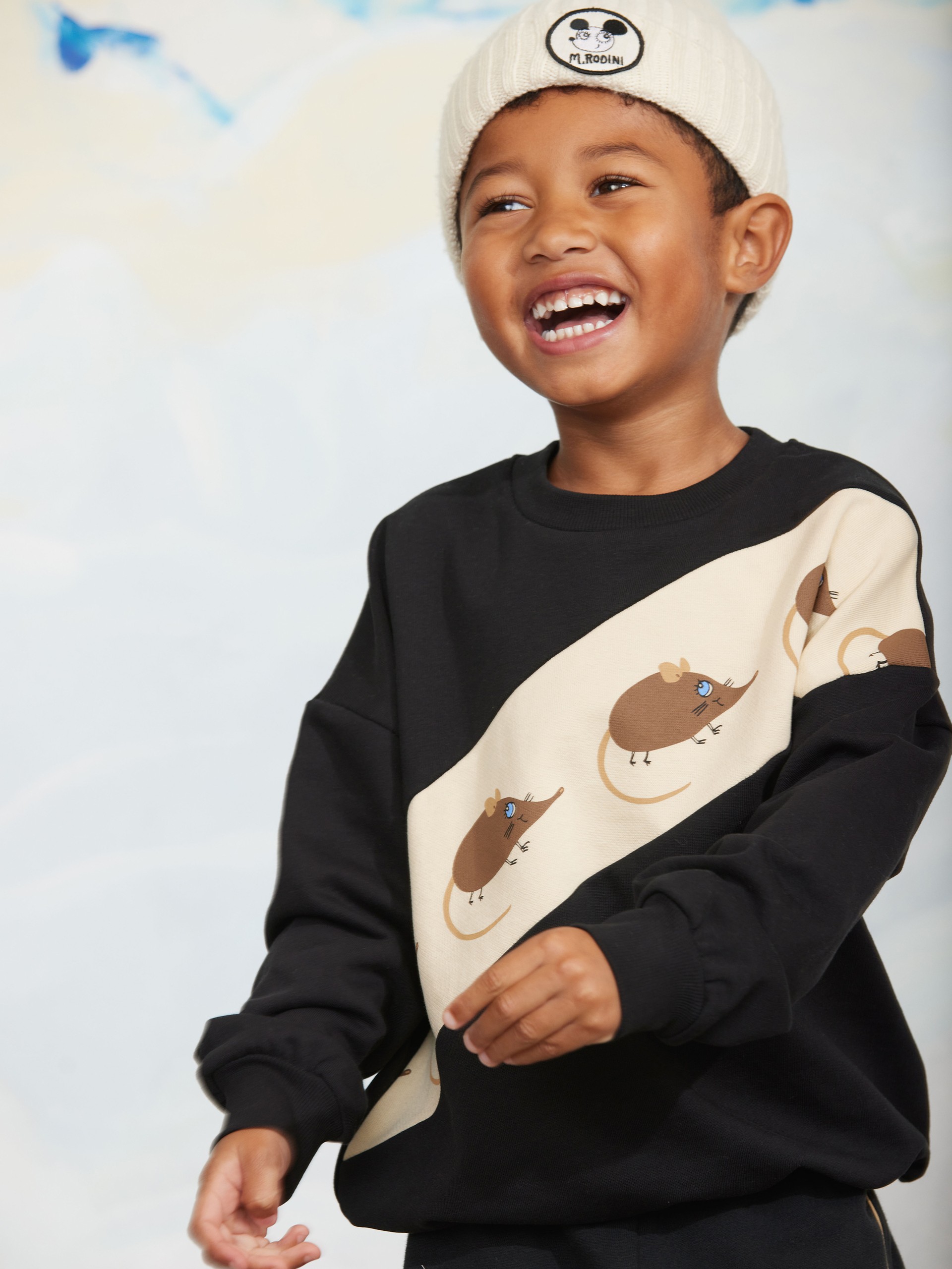 A smiling child wearing a white beanie and a black sweater with a beige stripe featuring cartoon mouse designs.