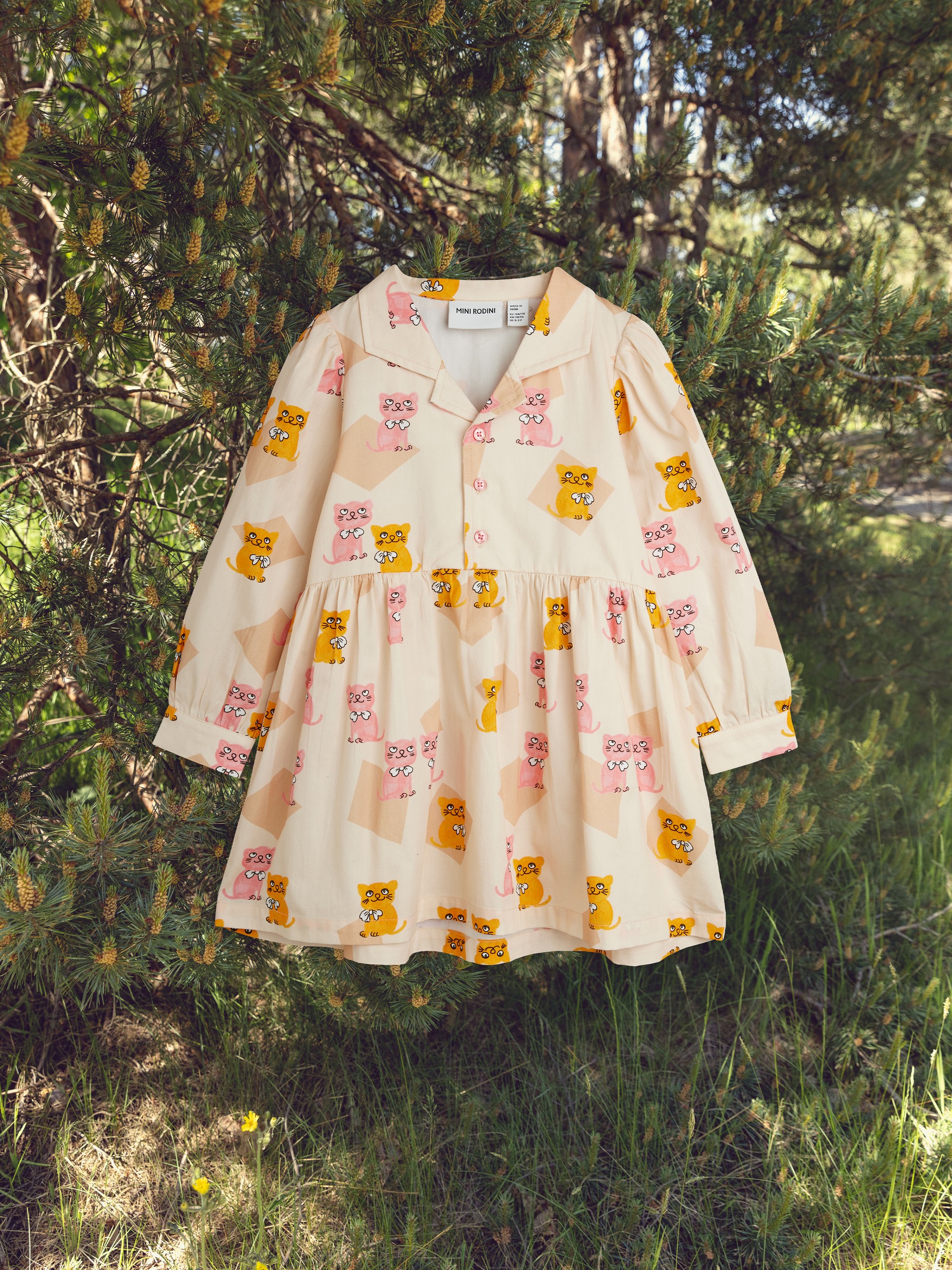 A child's dress with pink and yellow cartoon animal designs hangs on a tree branch in a sunlit forest setting.
