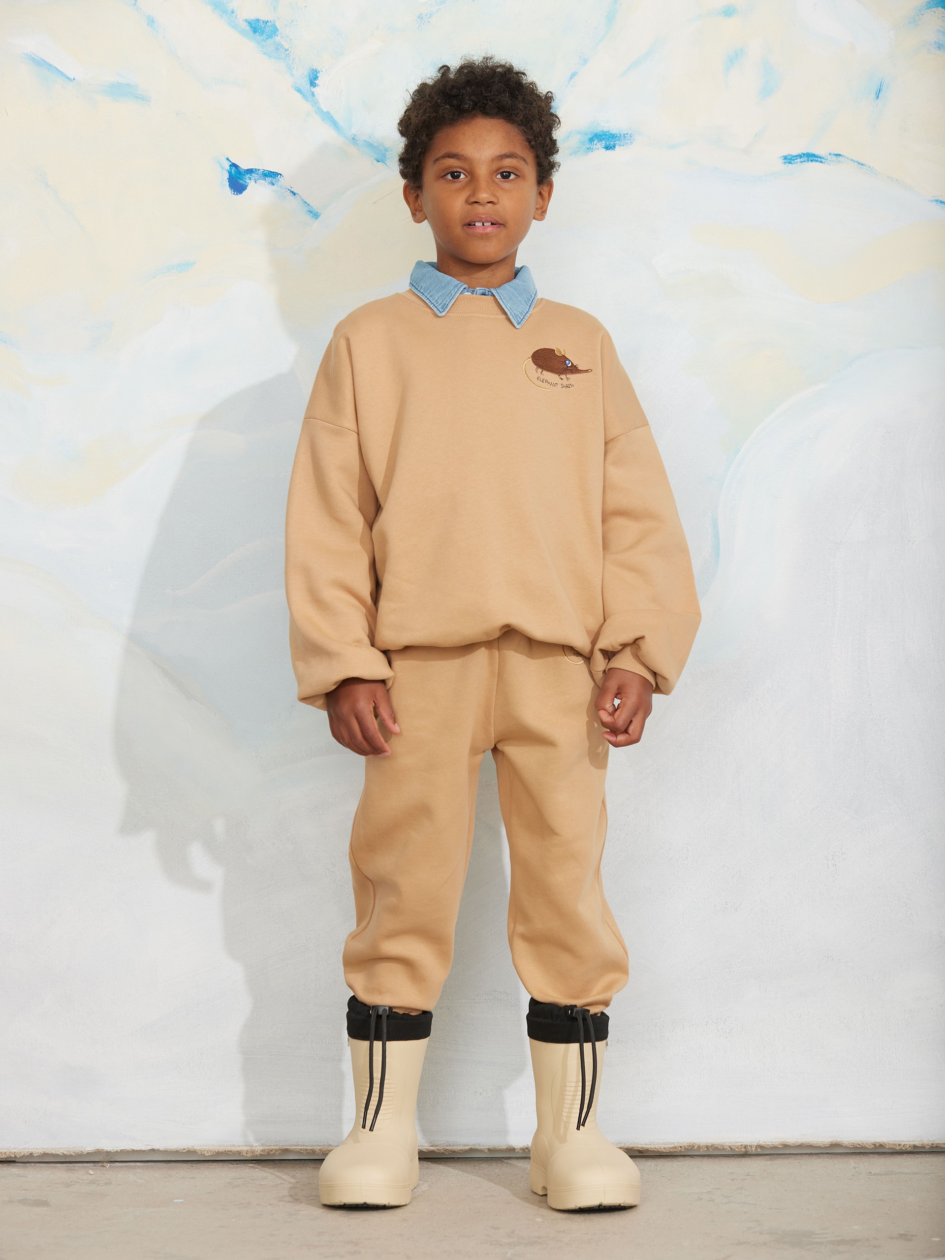 A child in a tan outfit with a blue collar stands against a light abstract background, wearing cream boots with black accents.