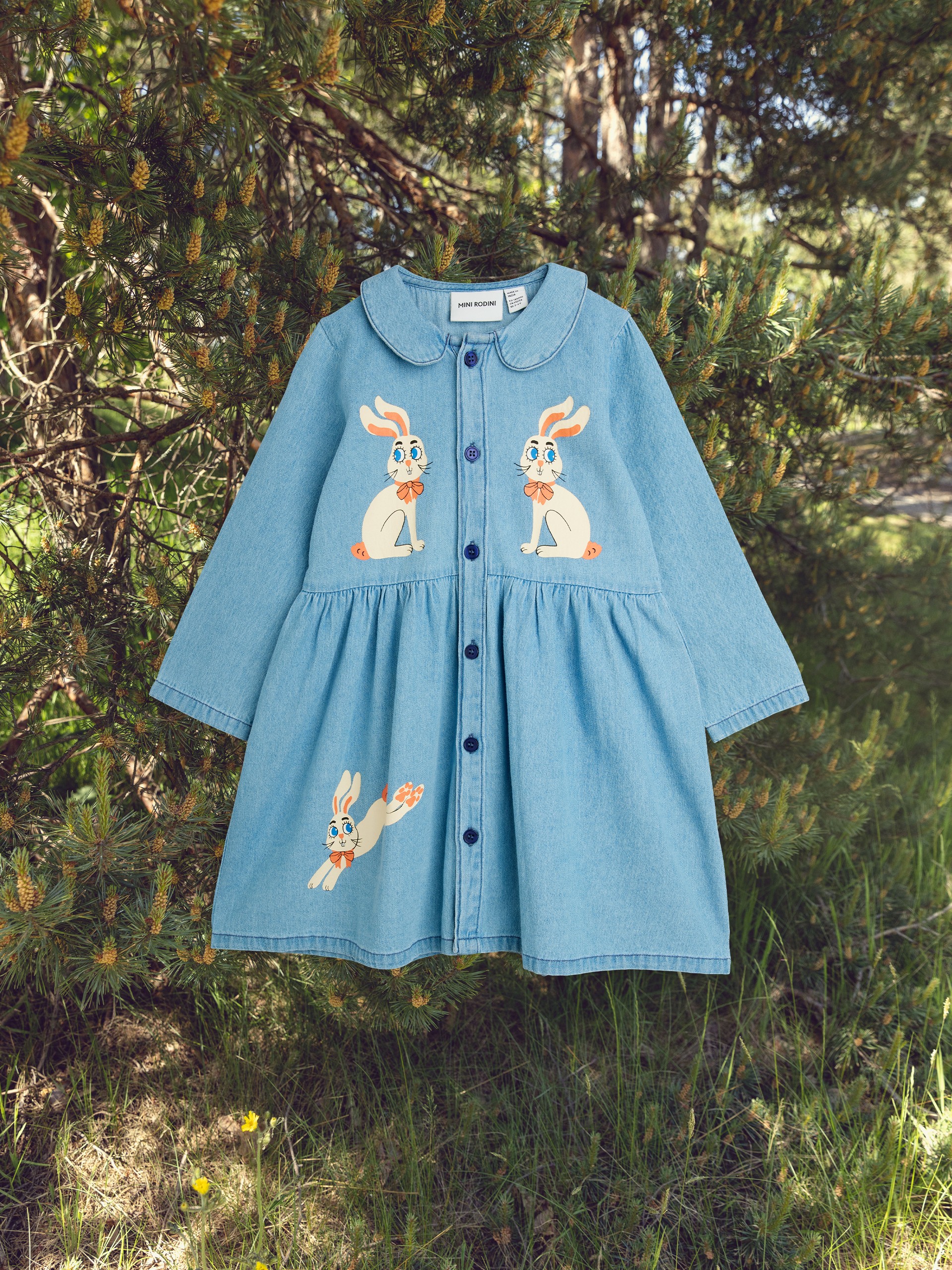 Children's denim dress with bunny designs, hanging on a tree branch in a sunny forest setting.