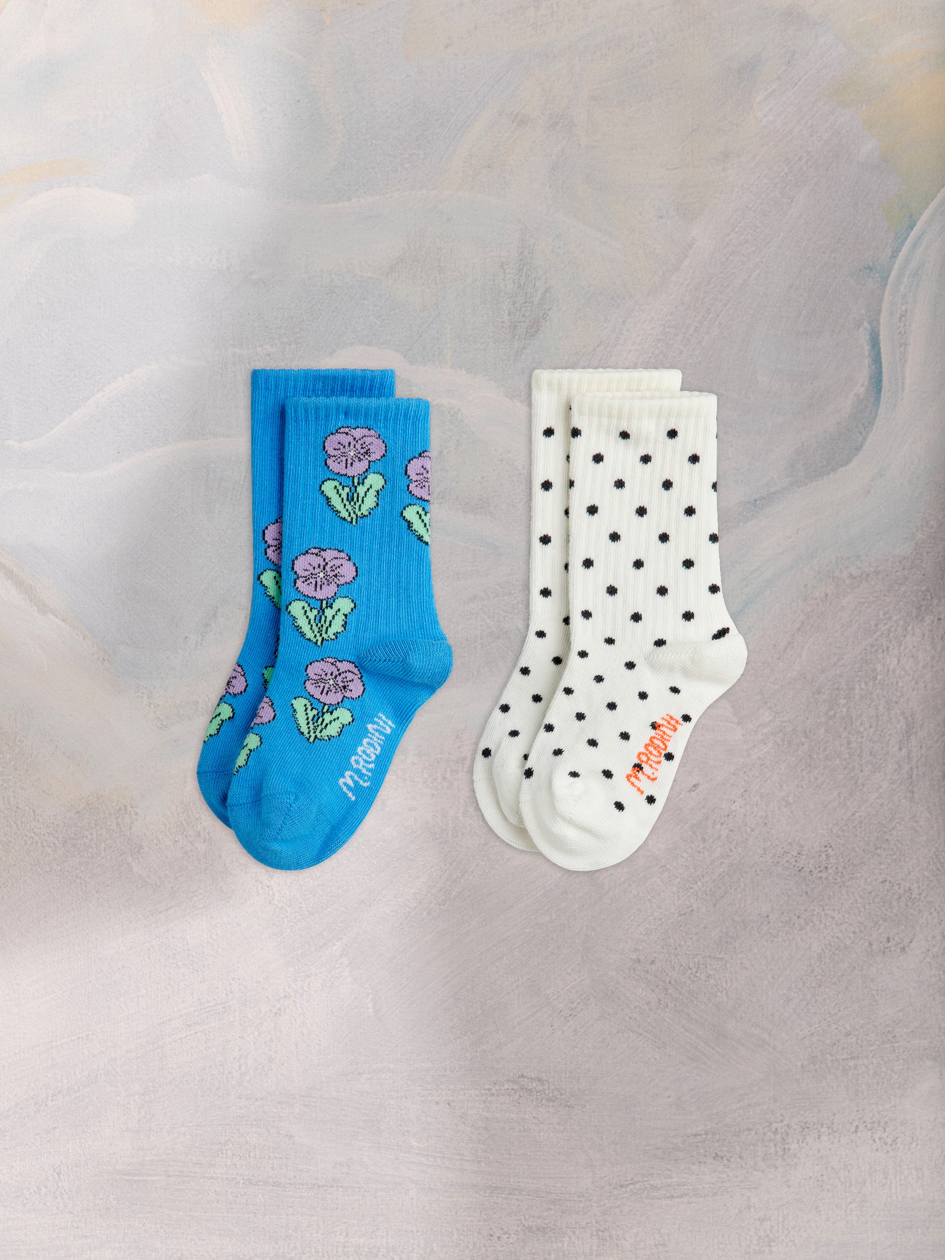 A pair of blue socks with purple flowers next to white socks with black polka dots, both labeled "MROOMI," on a textured gray background.