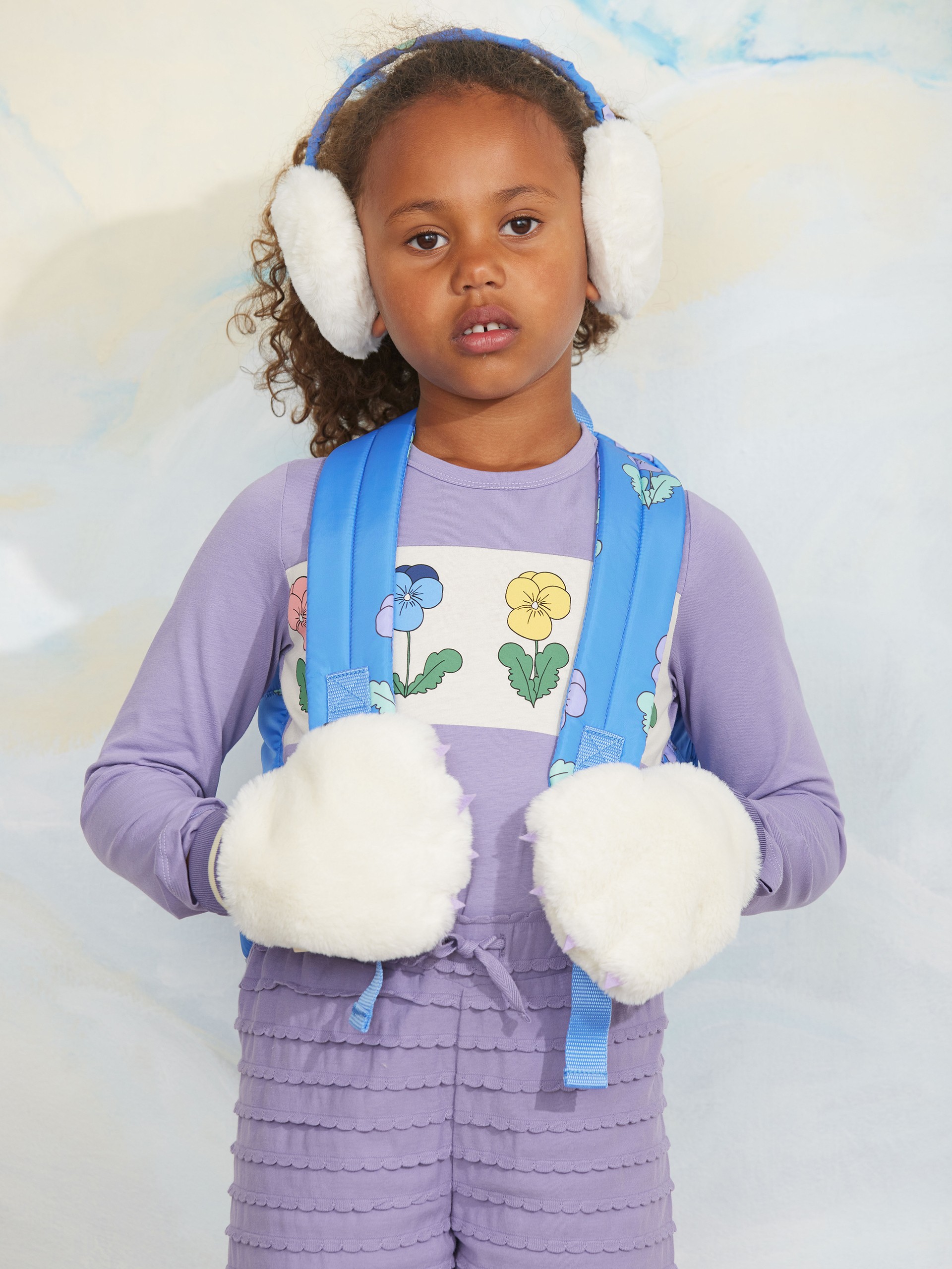 Child in a purple outfit with floral design, wearing blue earmuffs and fluffy white mittens against a light background.