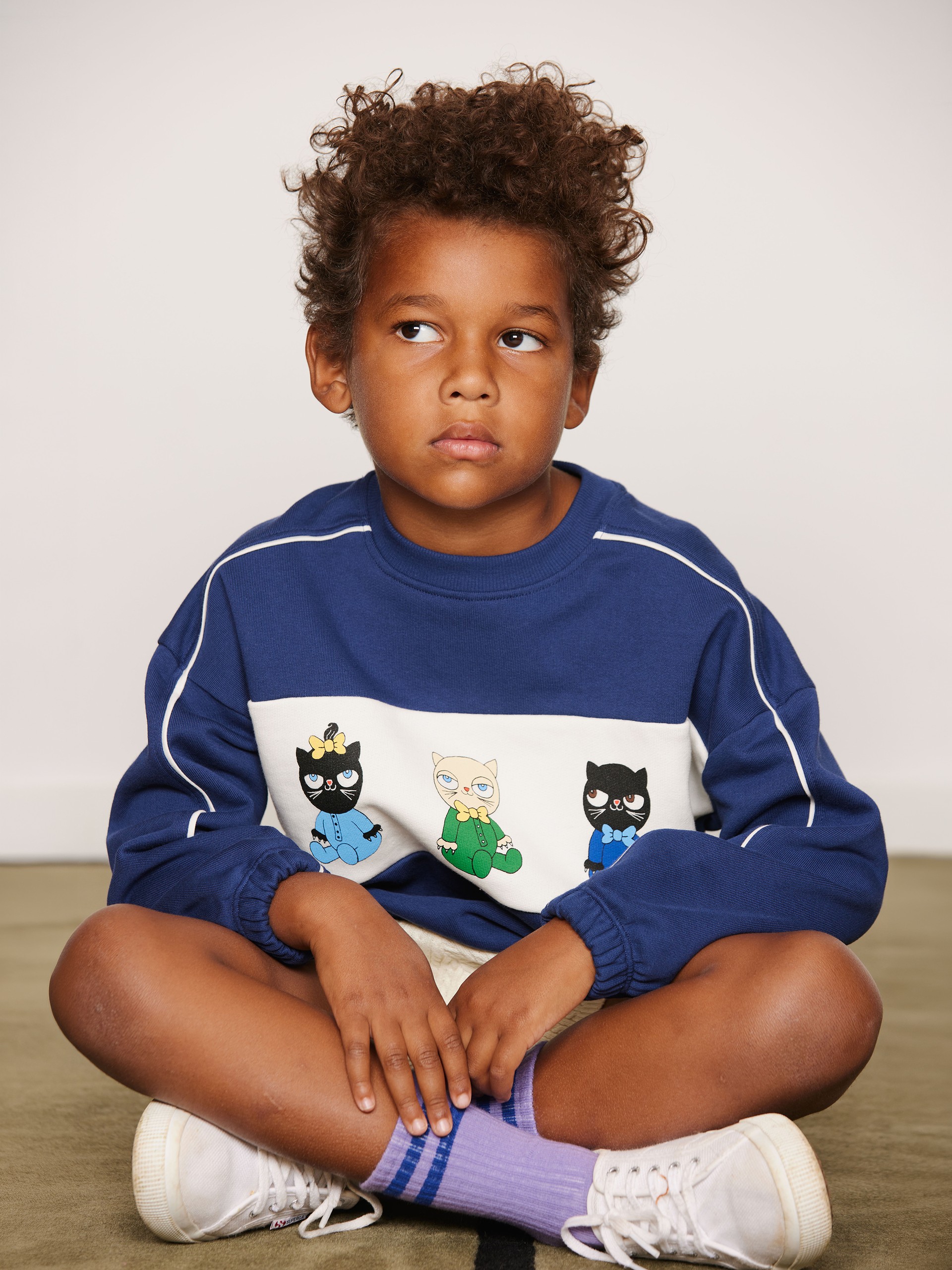 A child sits cross-legged, wearing a blue sweatshirt with Minibabies print from Mini Rodini, purple socks, and white shoes, looking contemplatively to the side.