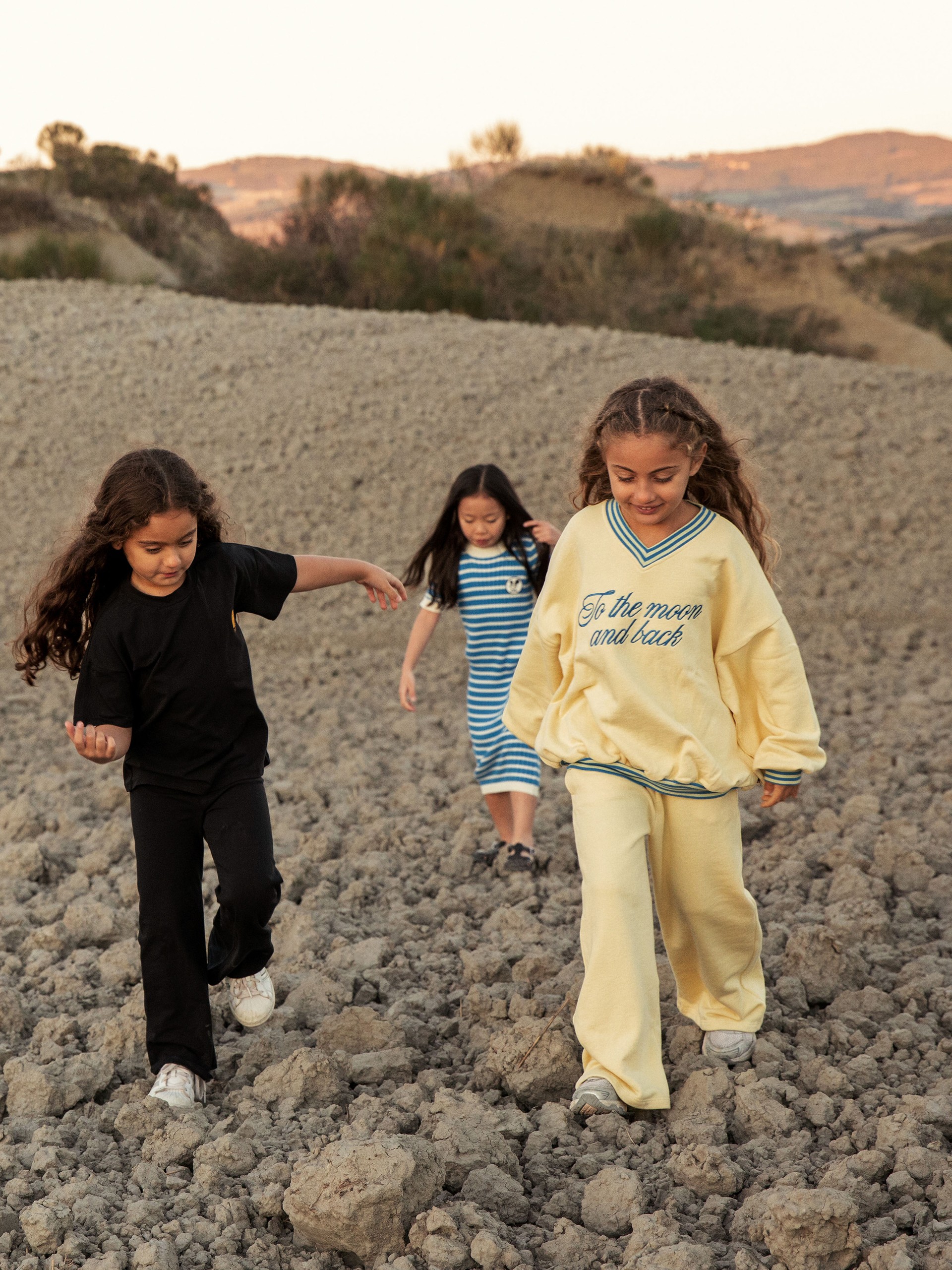 Three children joyfully walk on rocky terrain at sunset, wearing casual outfits from Mini Rodini Alien Affairs collection, with hills in the background.