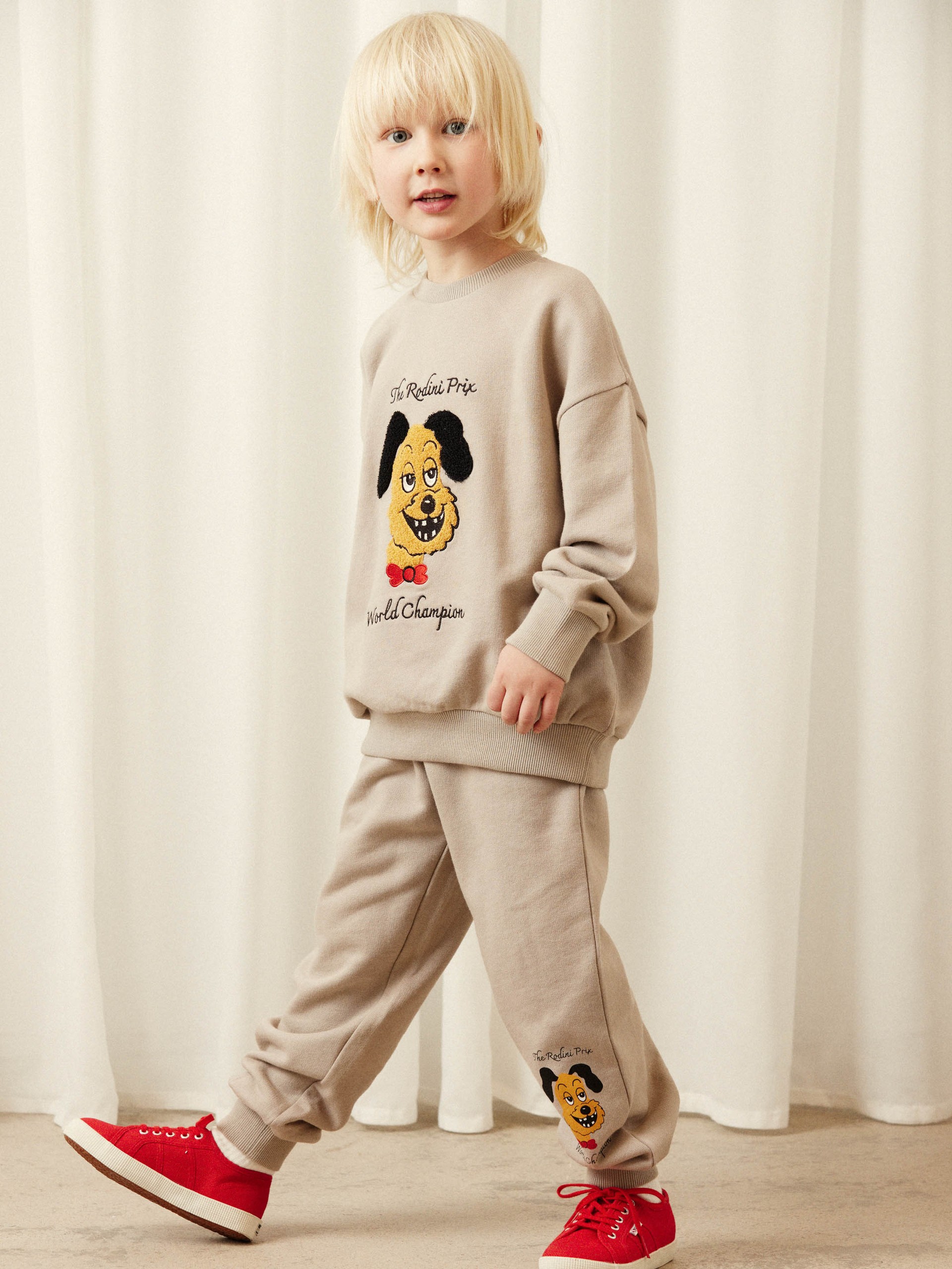 A child with blonde hair wearing a beige sweatsuit featuring a dog graphic and bright red sneakers, standing against a white curtain.