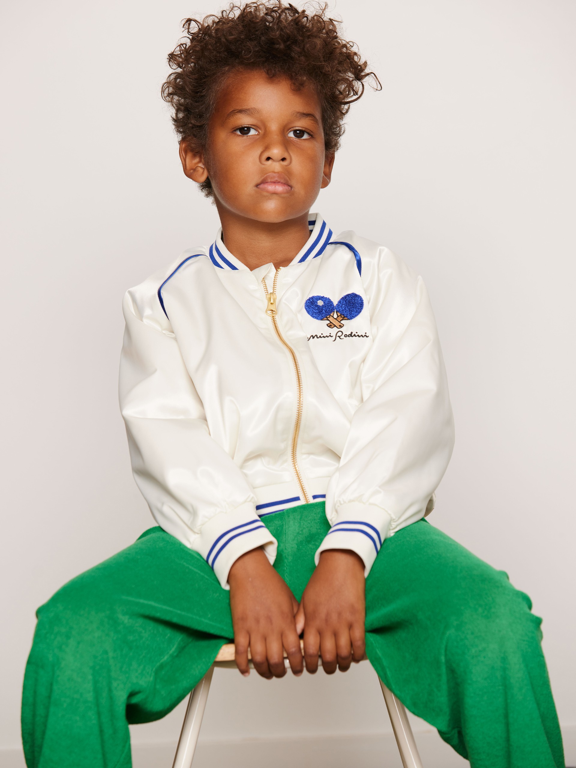 Child in a white jacket with blue details and green pants from Mini Rodini sits on a stool against a plain background.