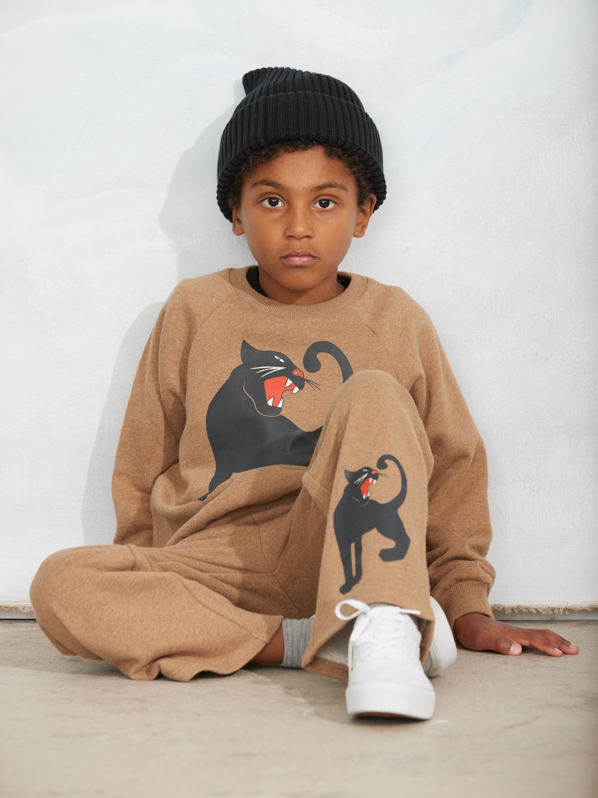 A child in a knit beanie sits against a wall, wearing a brown tracksuit featuring black panther graphics, and white sneakers.