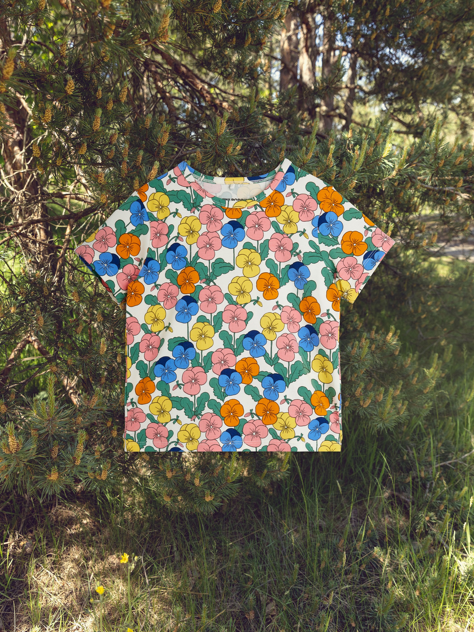 A colorful floral-patterned T-shirt hangs amidst green pine branches in a sunny, natural setting.