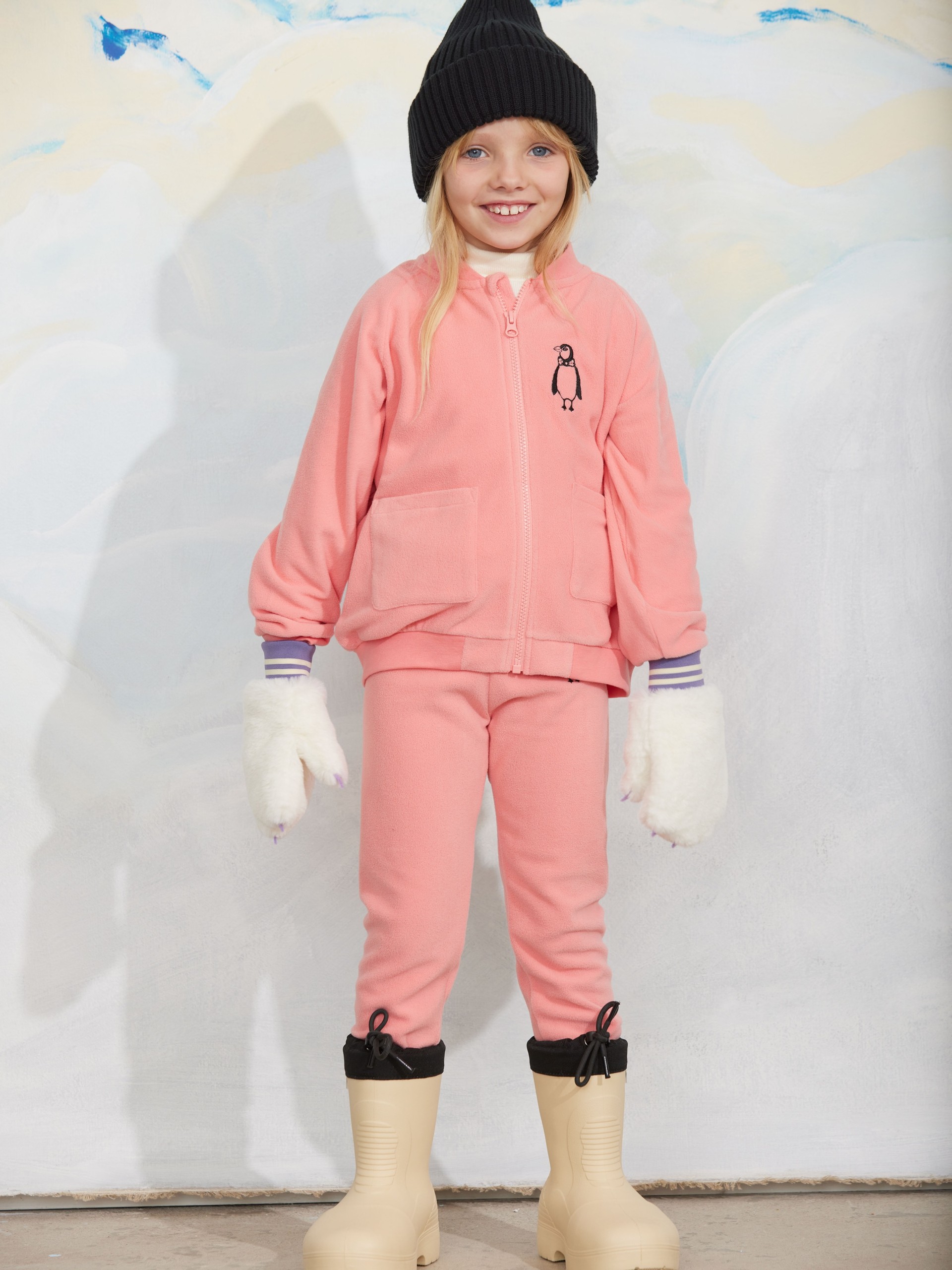 A smiling child wearing a pink outfit with penguin embroidery, black beanie, white mittens, and beige boots stands against a snowy background.