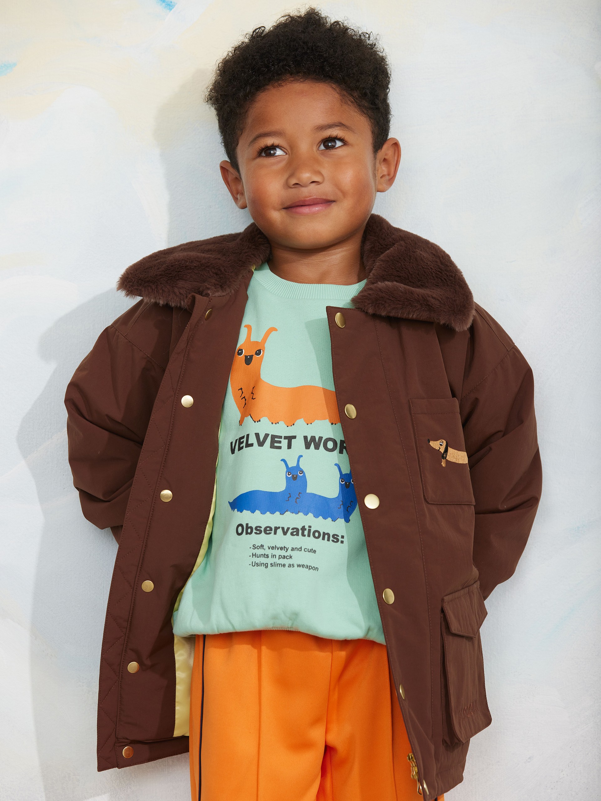 A child wearing a brown jacket with a fur collar, light blue shirt with cartoon slugs, and orange pants, smiling against a light background.