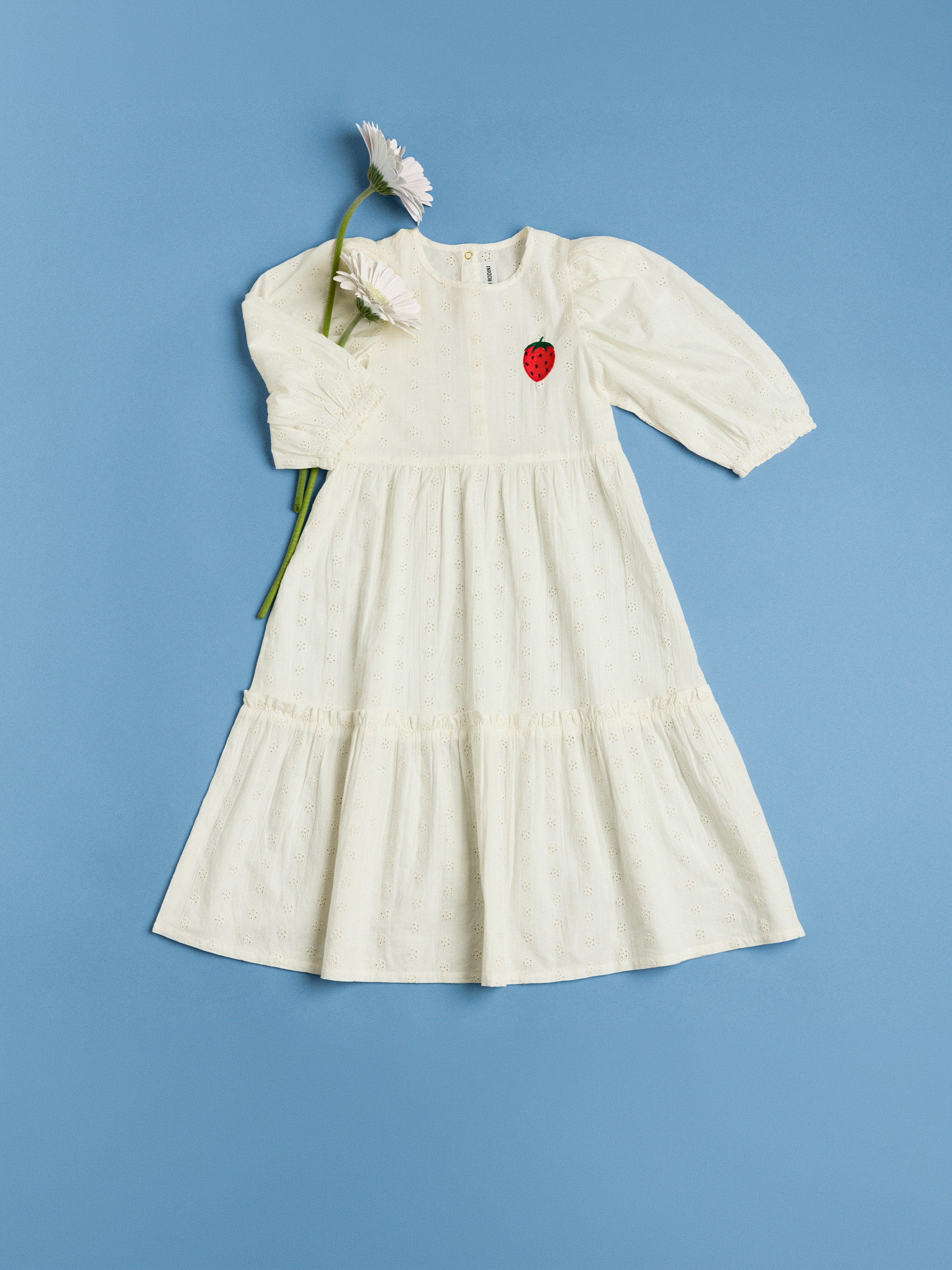 Cream dress from Mini Rodini with puffed sleeves and a strawberry patch, adorned with white flowers, on a blue background.
