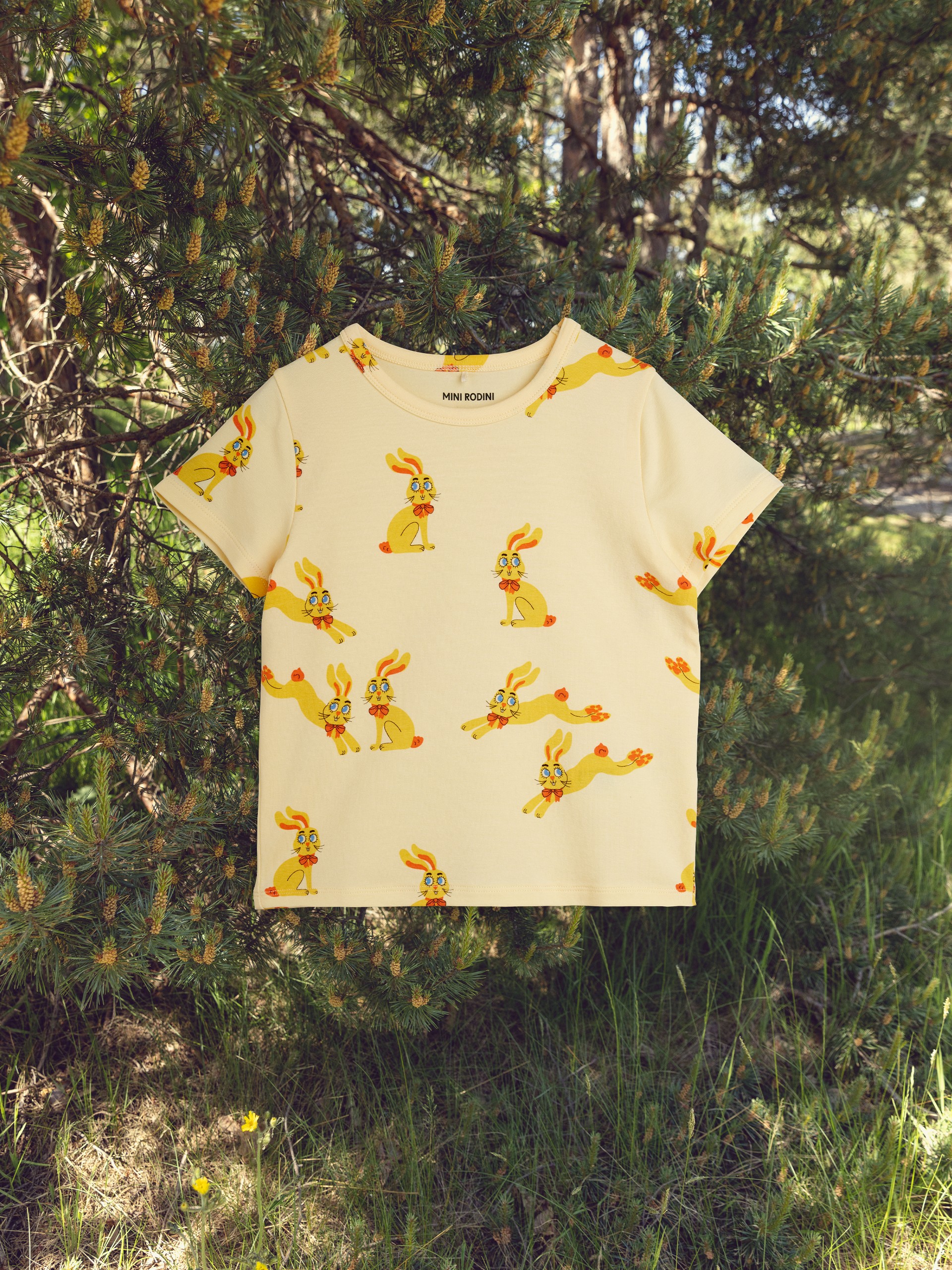 A children's shirt with a yellow rabbit pattern is hanging outdoors among green pine branches.