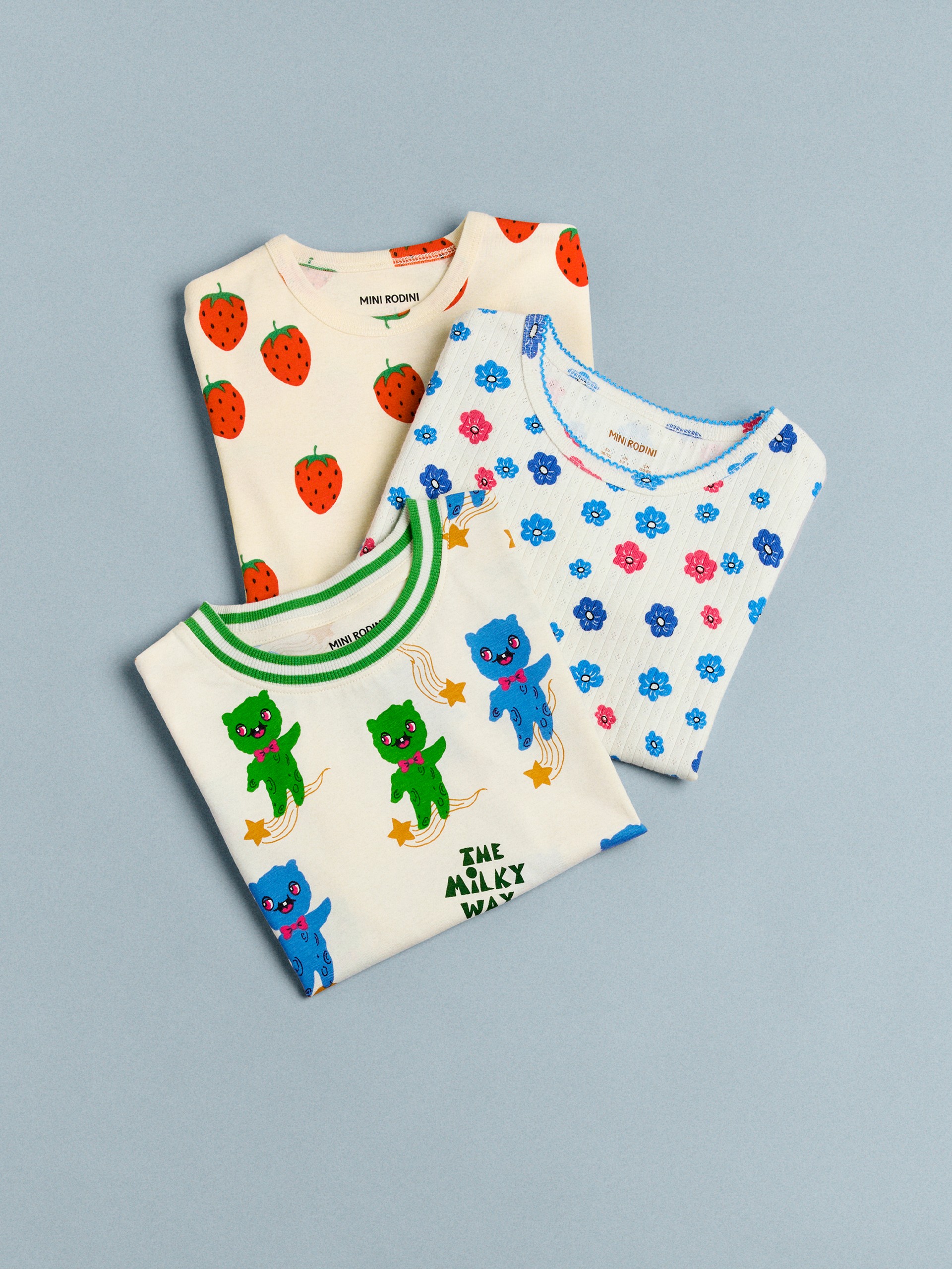 Three folded T-shirts from Mini Rodini on a blue background: one with strawberries, one with blue and pink flowers, and one with colorful Aliens and stars.