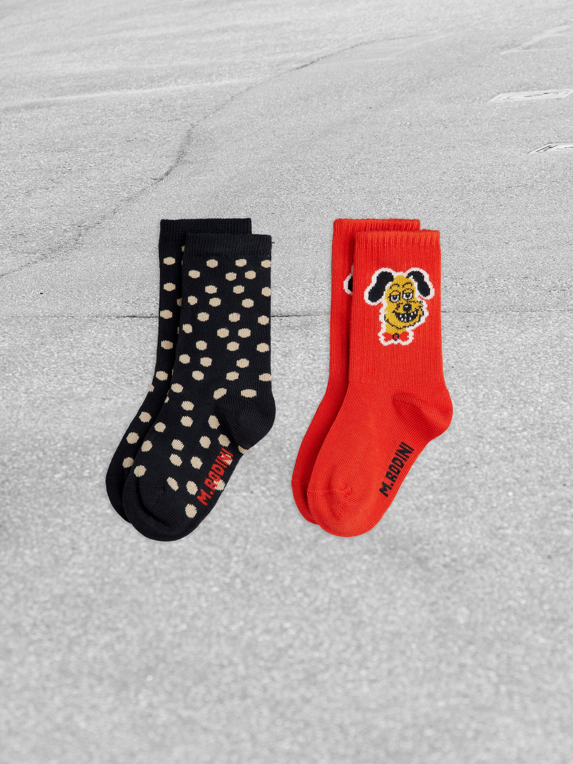 Two pairs of socks on a gray surface: one black with beige dots, the other red featuring a cartoon dog's face.