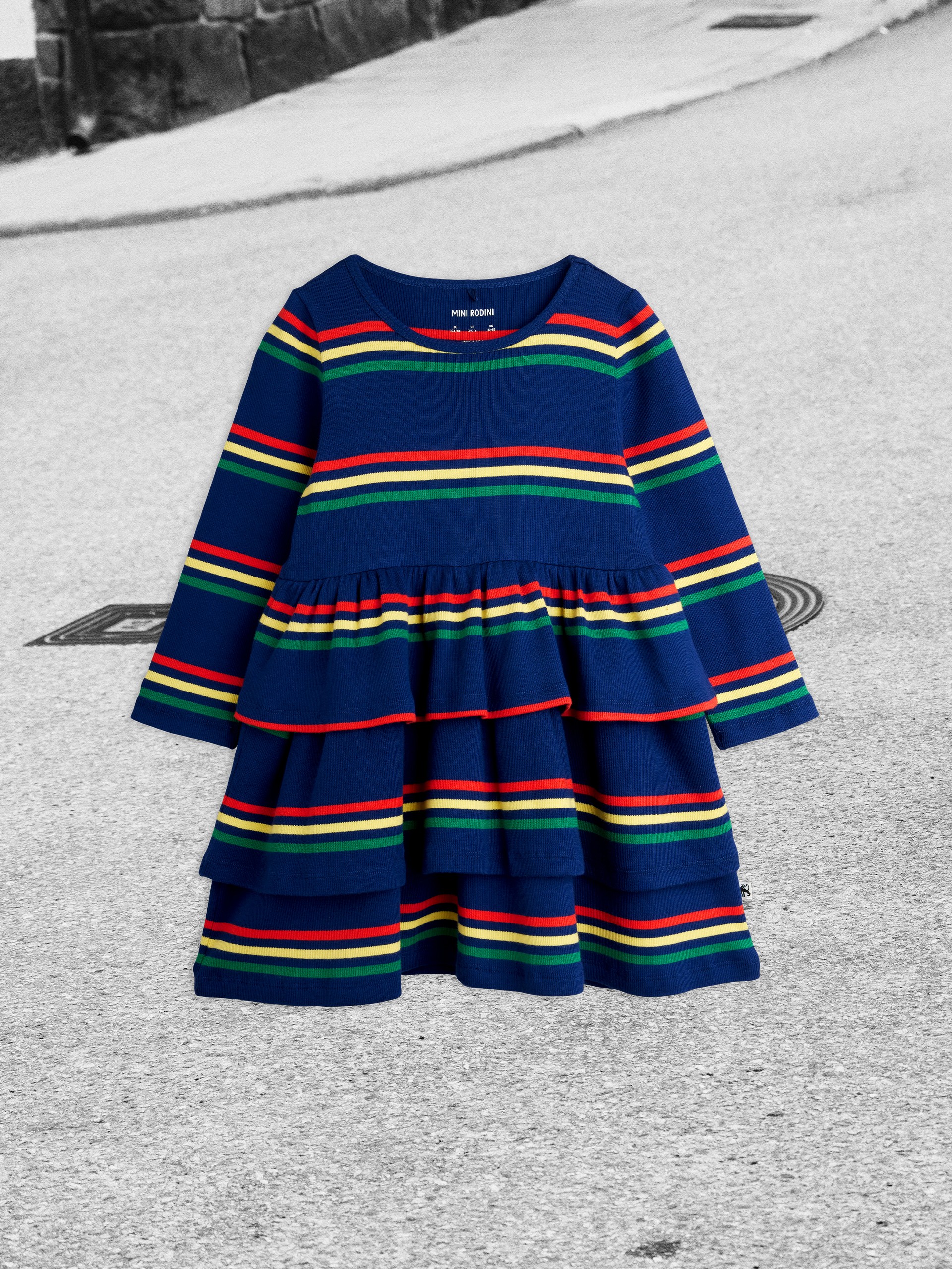 A colorful striped blue dress with long sleeves and tiered layers displayed on a grey concrete background.