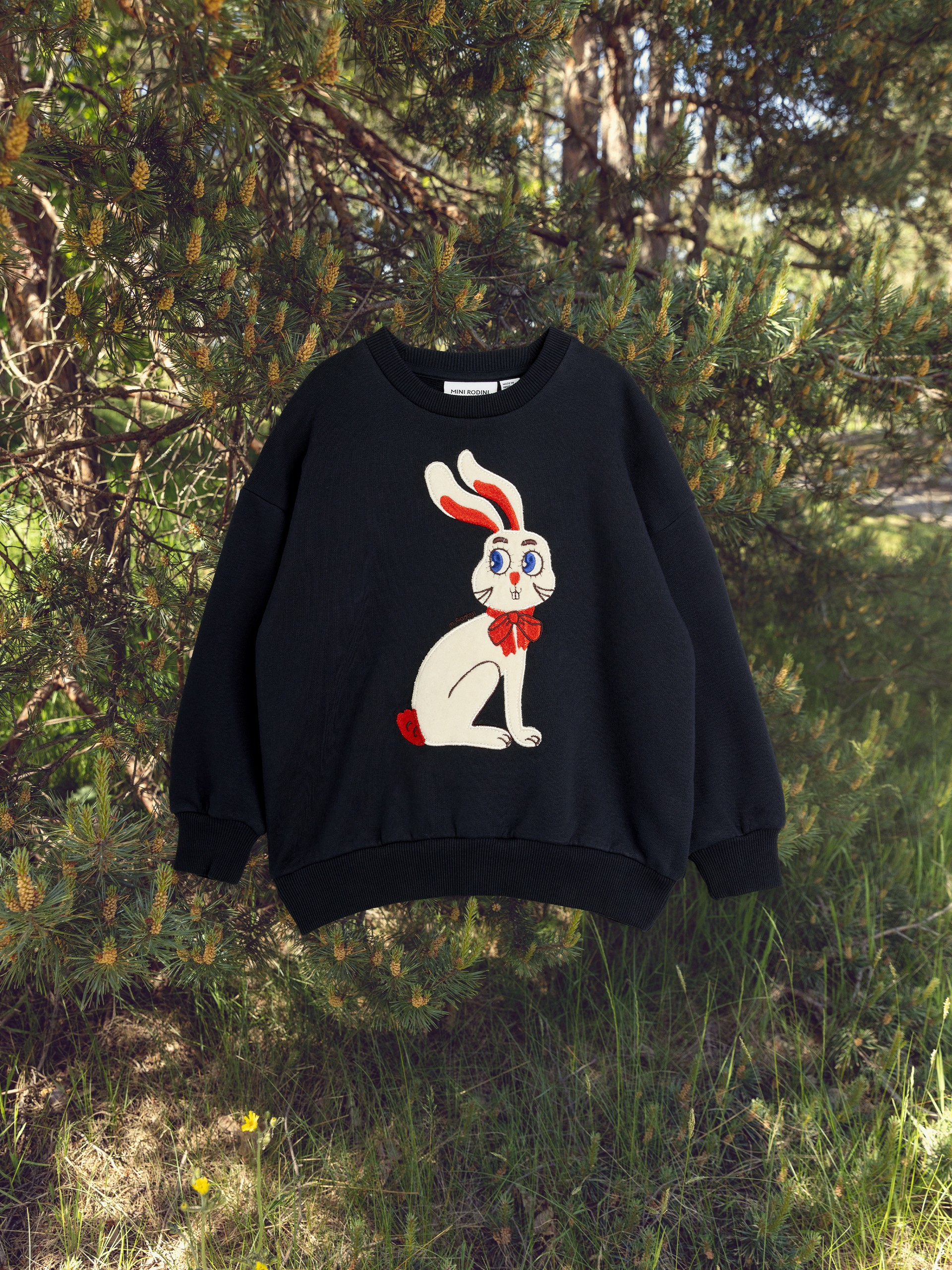 A black sweater featuring a cartoon bunny with red ears and bow is displayed outdoors with green pine trees and grass in the background.
