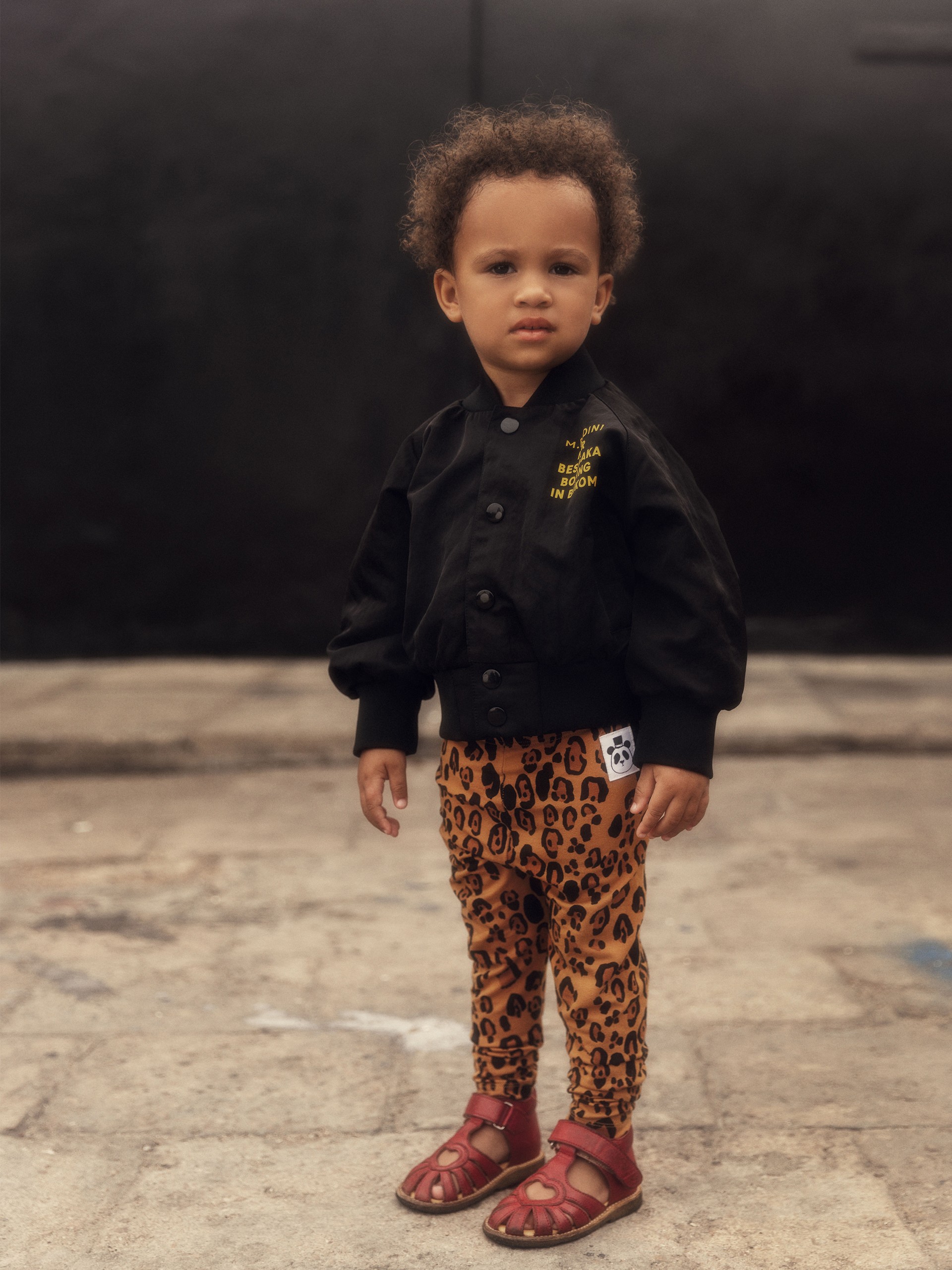 Young child in Besesaka baseball jacket and basic leopard leggings from Mini Rodini, and red sandals standing on a concrete surface with a neutral expression.