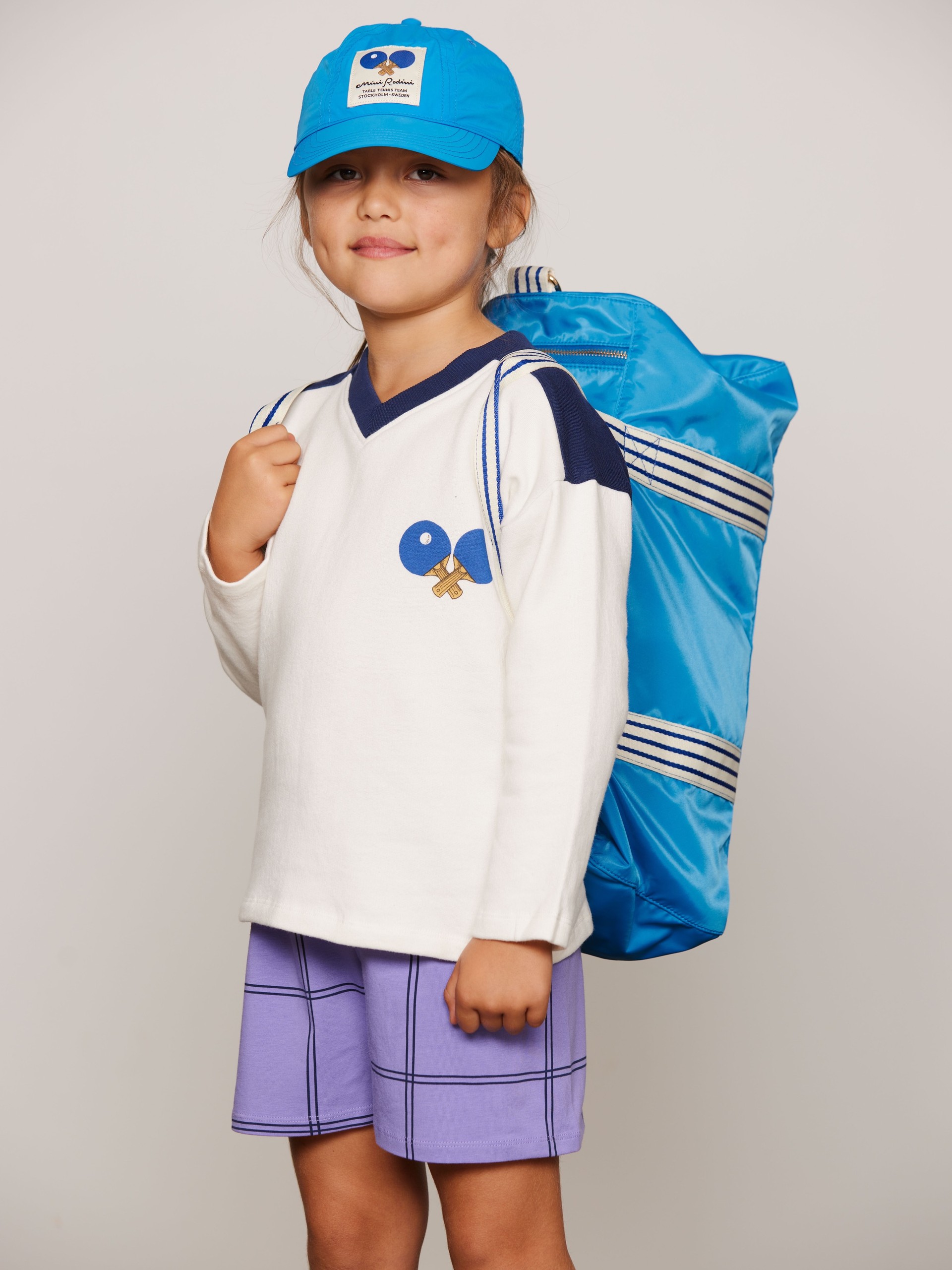 Child wearing Table tennis patch nylon cap and Table tennis gym bag from Mini Rodini, white sweater with ping pong paddles, and purple shorts, all from Mini Rodini.