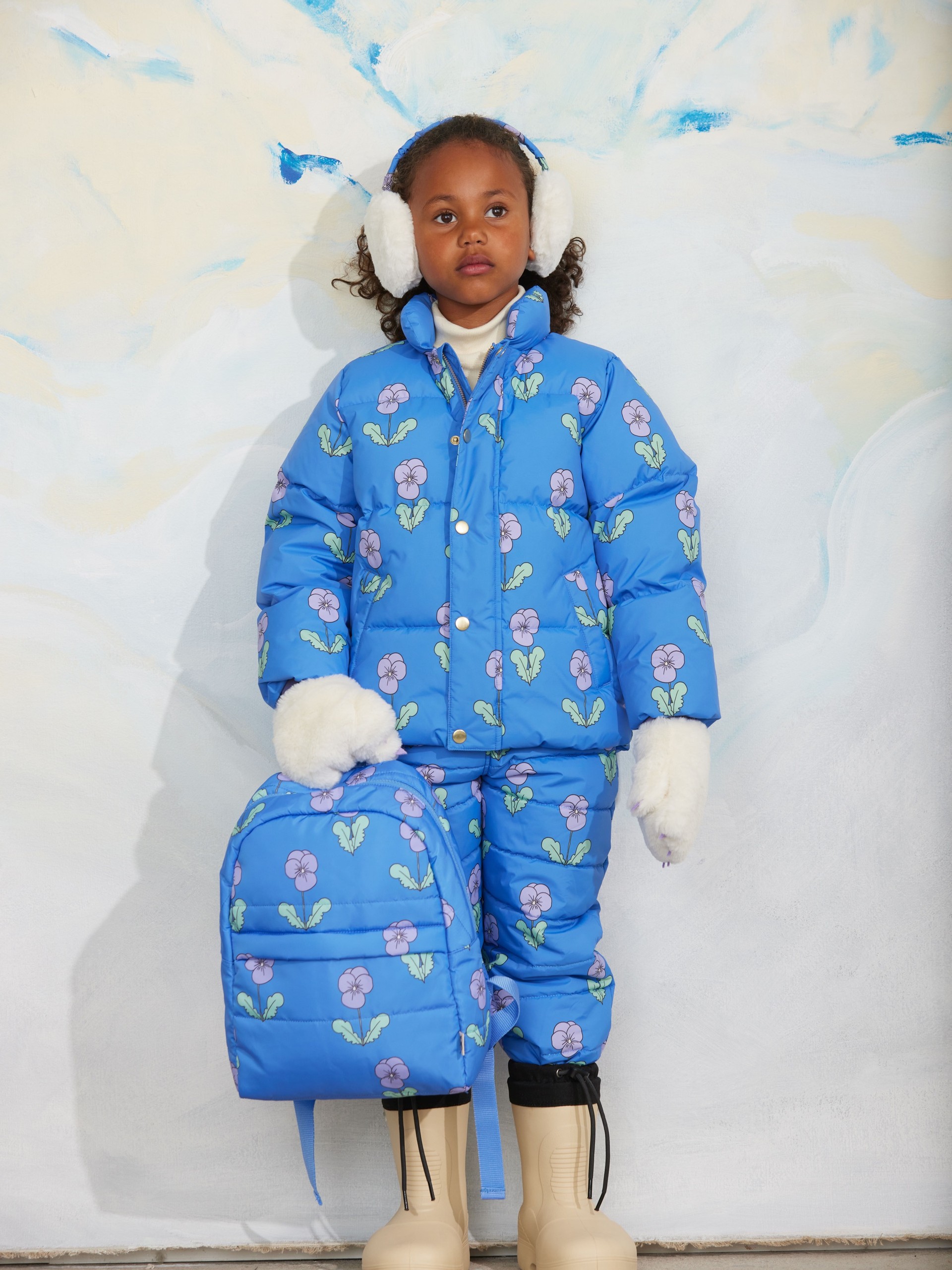 Child wearing blue floral-patterned snowsuit, earmuffs, and gloves, holding a matching backpack, stands against a snowy backdrop.