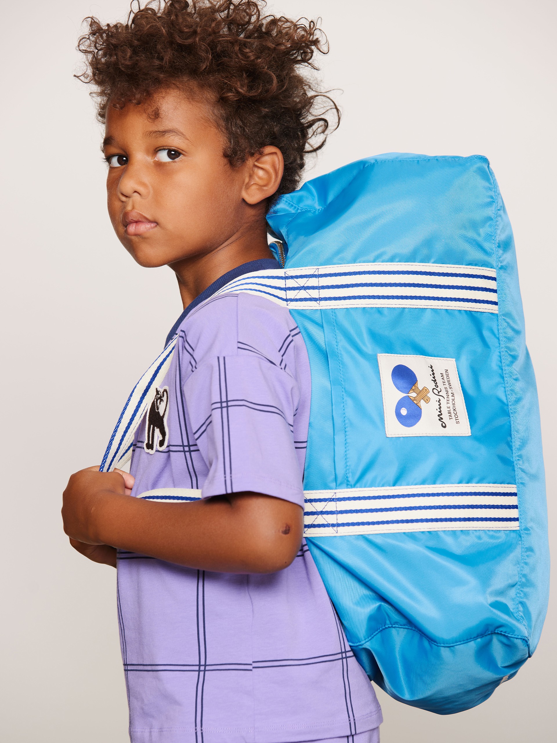 Young boy with curly hair in a purple checkered shirt, carrying a large blue backpack, looking to the side.
