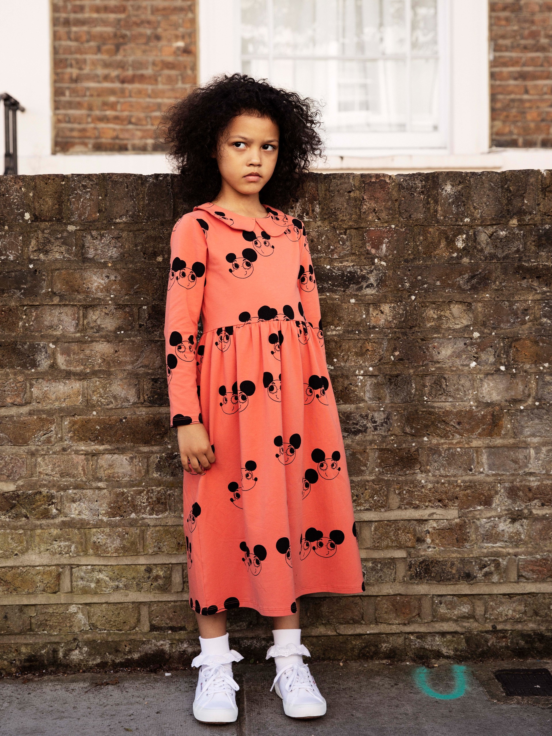 A young girl with curly hair stands against a brick wall, wearing a peach dress with black cartoon faces and white sneakers.