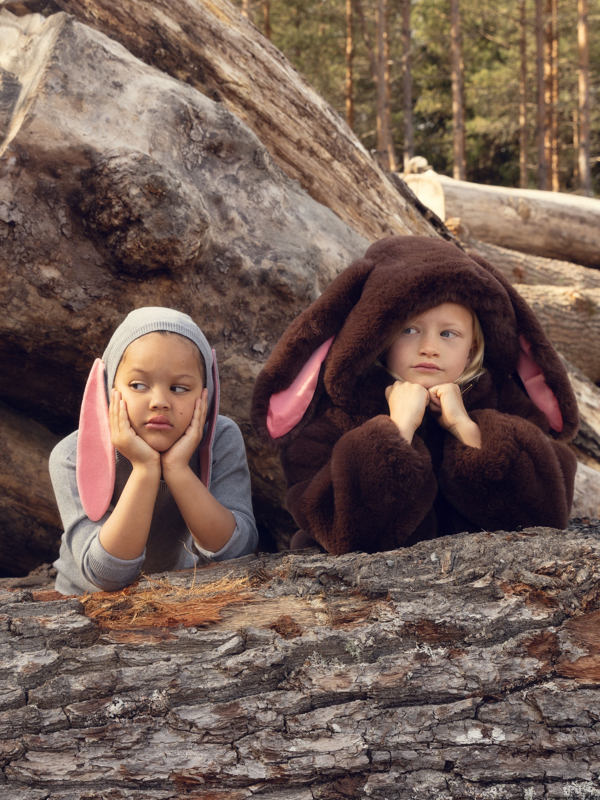 Two children in bunny costumes rest their chins on a log in a forest, looking thoughtful and curious.