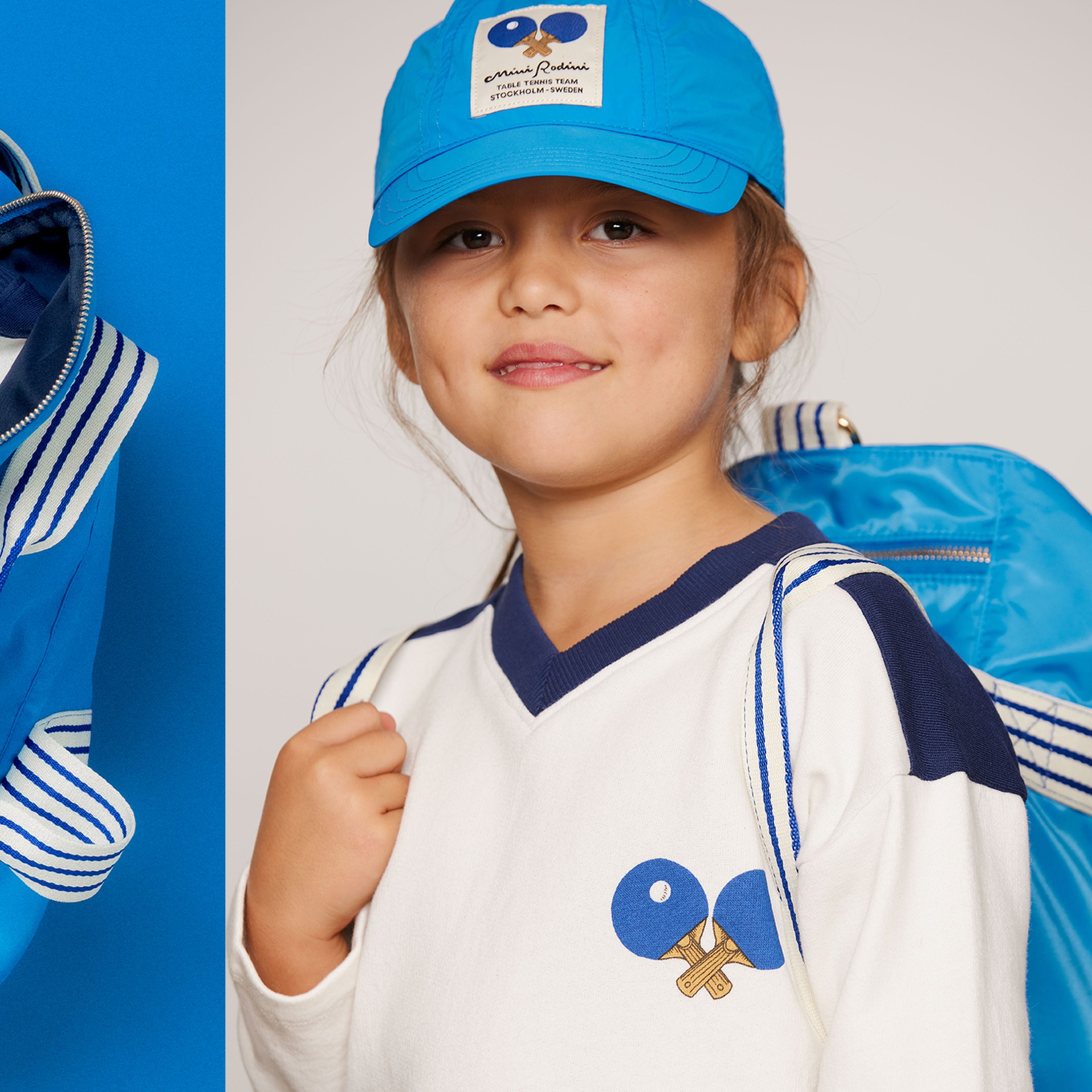 Table tennis gym bag from Mini Rodini with a water bottle, white shirt, Rubik's Cube, headphones, and sneakers on a blue background.