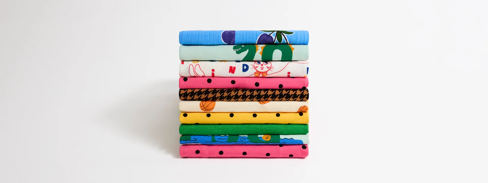A neatly stacked pile of colorful clothes from Mini Rodini Upcycling collection with various patterns, including Polka dots, Loch ness, Basketball print and vibrant designs, on a white background.