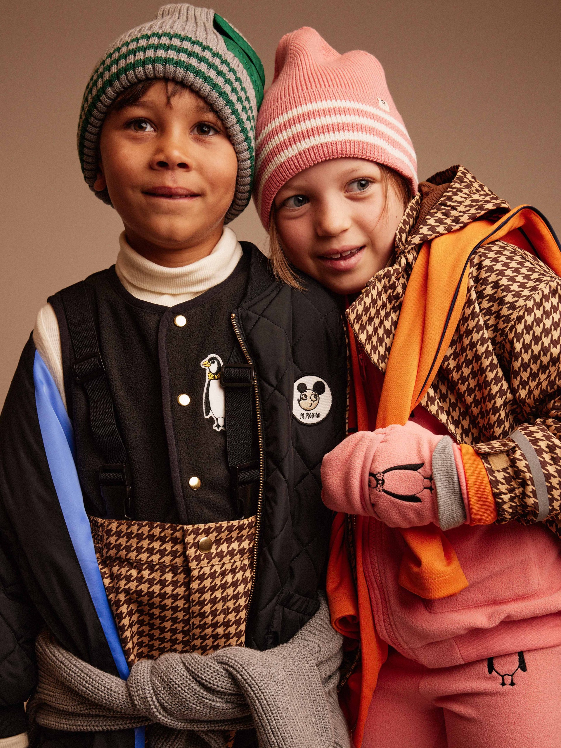 Two children wearing winter hats and houndstooth patterned clothing, standing closely together and smiling. One wears pink, the other wears brown and blue.
