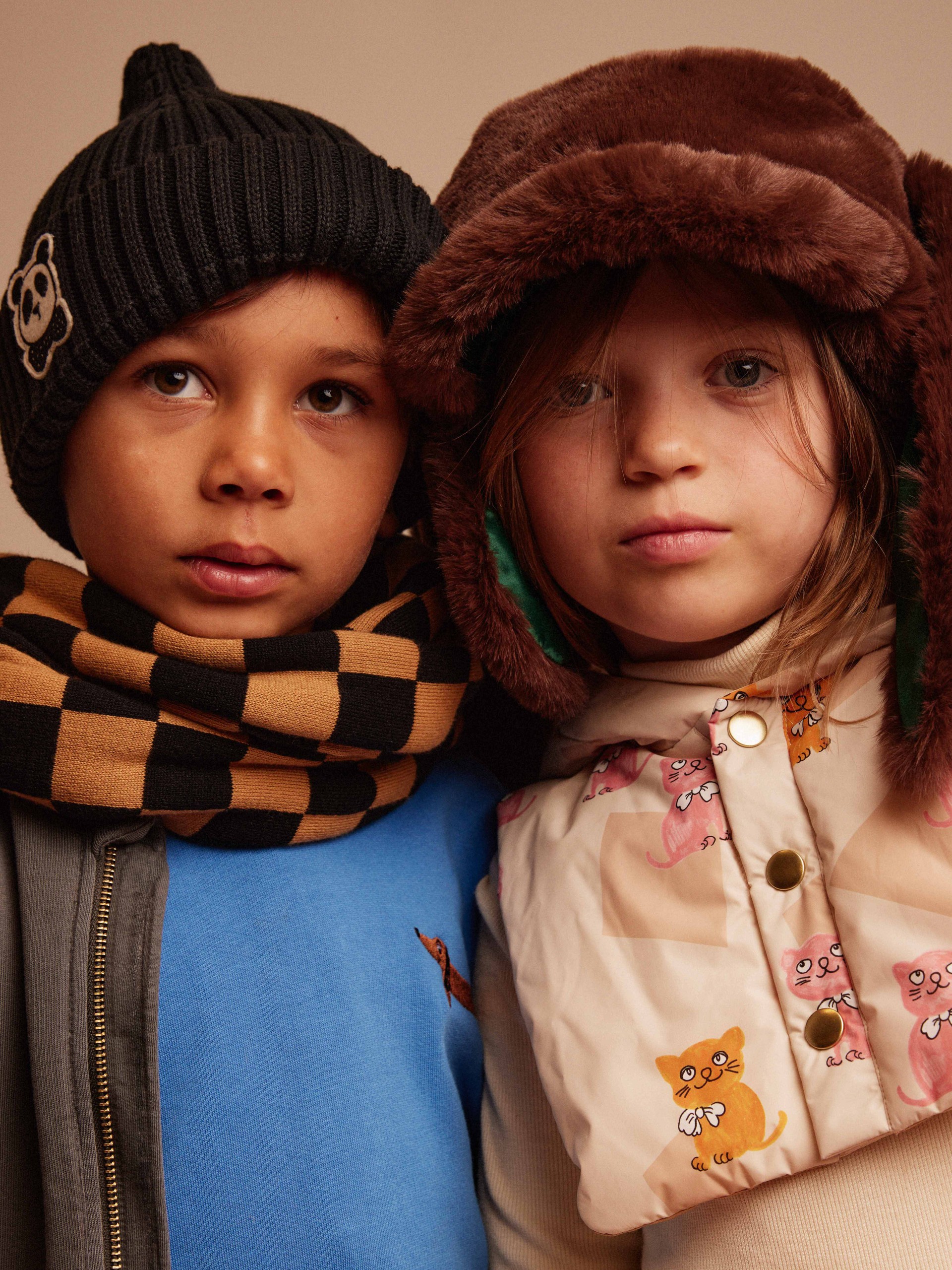 Two children wearing winter hats and scarves, standing side by side. One wears a panda-beanie and the other a furry hat with ear flaps.