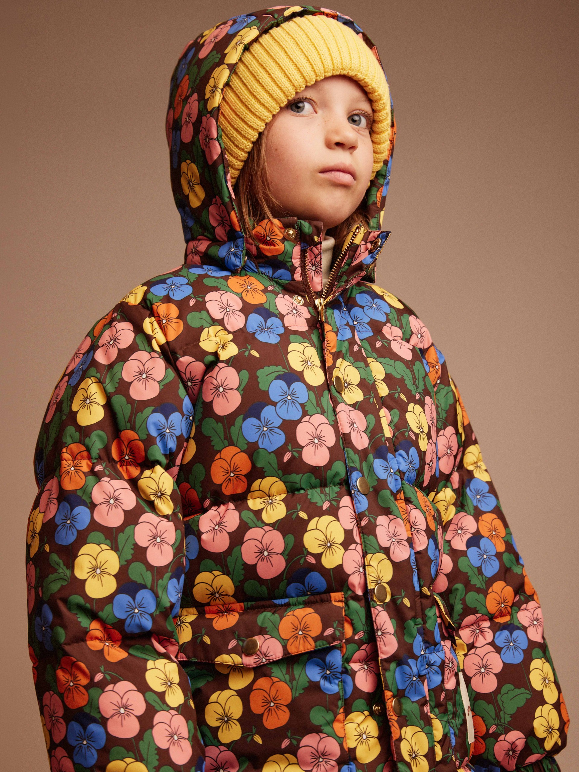 Child wearing a vibrant floral-patterned puffer jacket and a yellow knit beanie, looking to the side against a neutral background.