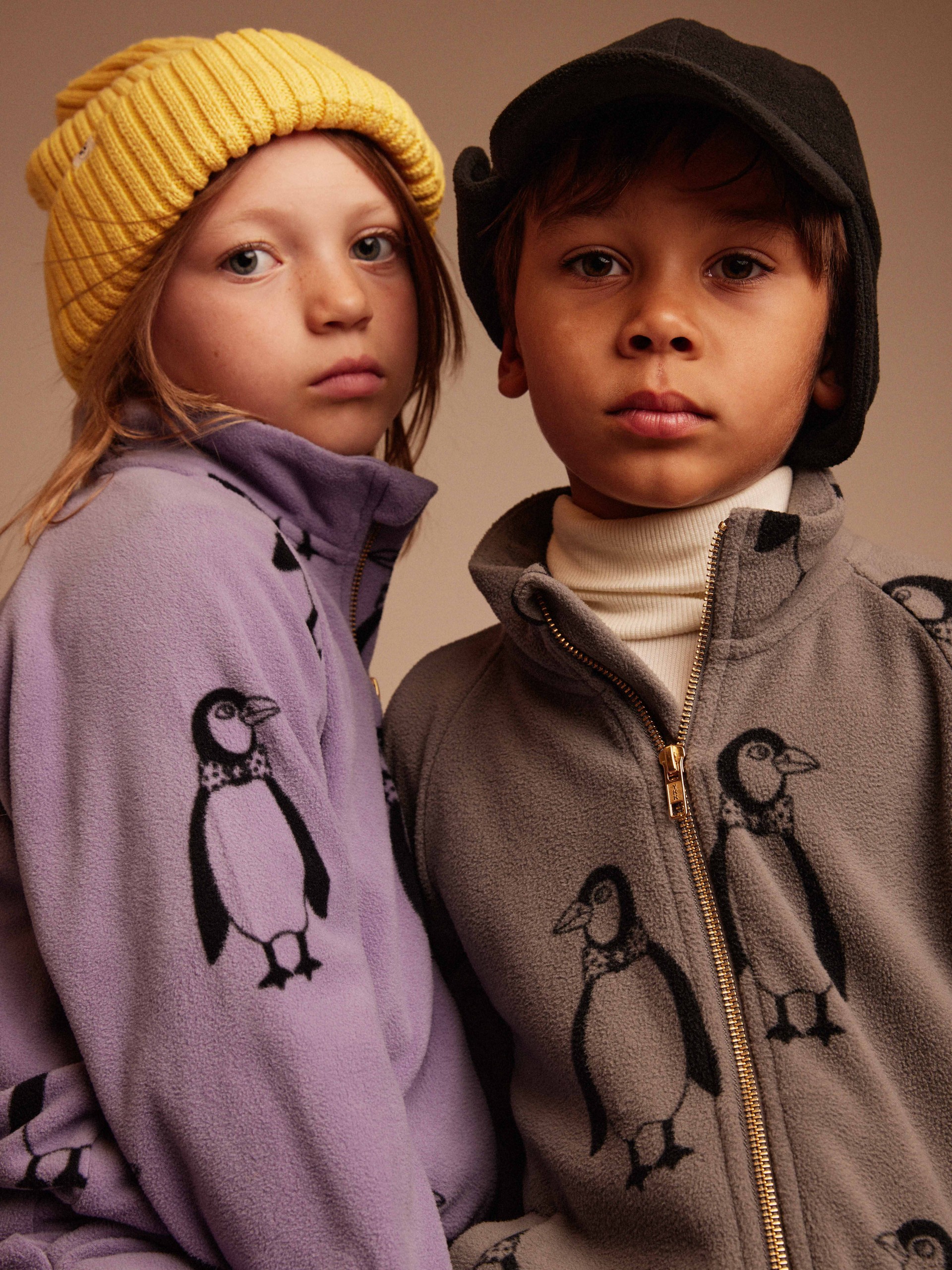 Two children looking at the camera, wearing fleece jackets with penguin patterns. One child wears a yellow beanie, the other a black cap.