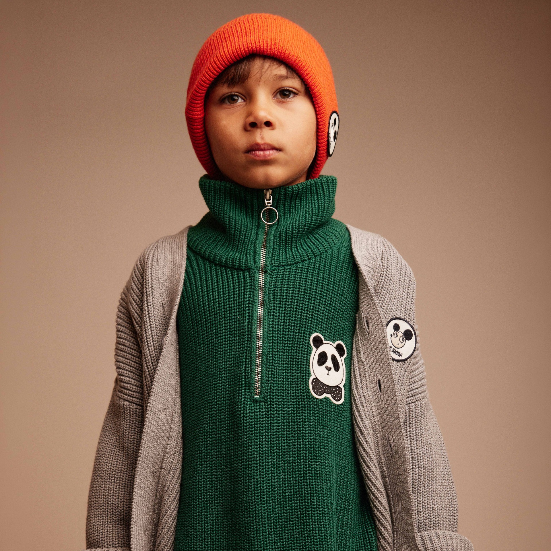 A child wears a green sweater with a panda patch, a gray cardigan, beige pants, and an orange beanie with a panda emblem, posing against a plain background.