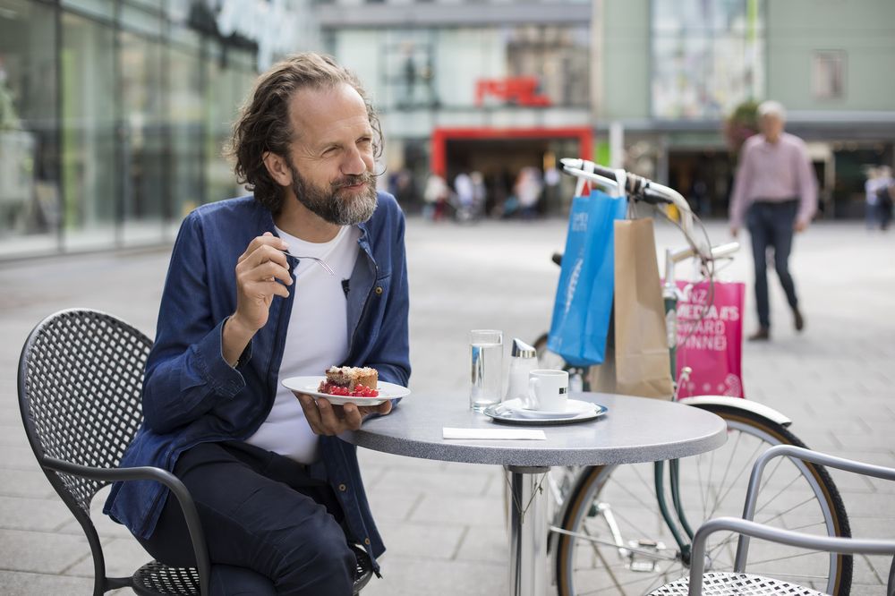 Ein Mann sitzt draußen an einem Tisch und isst ein Stück Kuchen. Hinter ihm ist ein Fahrrad mit mehreren Einkaufstüten am Lenker zu sehen.,