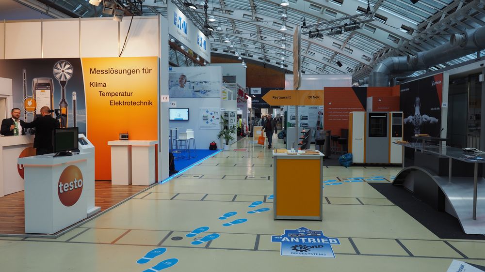 Eine großzügige Ausstellungshalle mit verschiedenen Ständen, Bannern und blauen Markierungen auf dem Boden, in der die Besucher die Ausstellungsstücke betrachten.