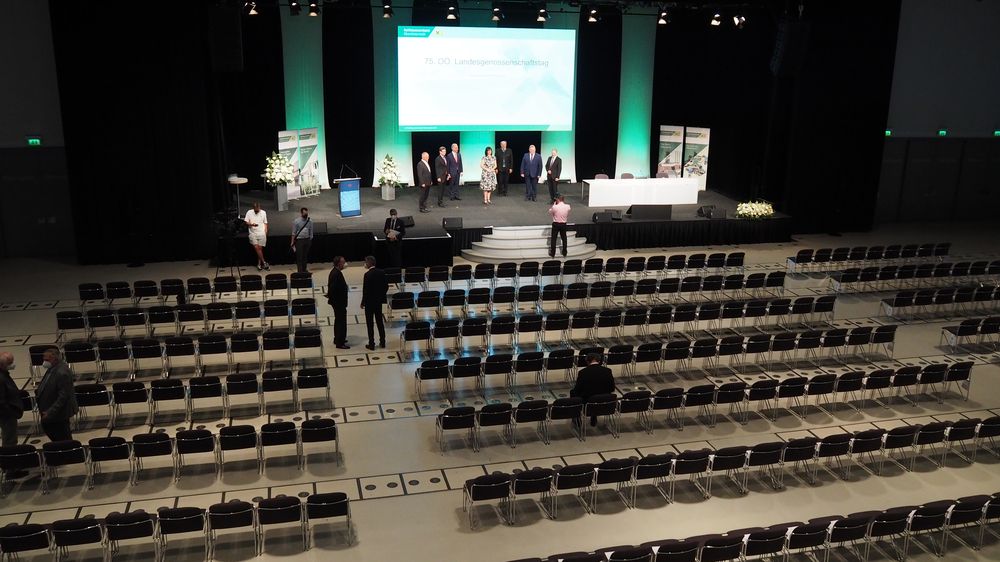 Ein großer Konferenzsaal mit Reihen leerer Stühle, einer Bühne mit Leinwand und einigen wenigen Menschen, die herumstehen und umhergehen.