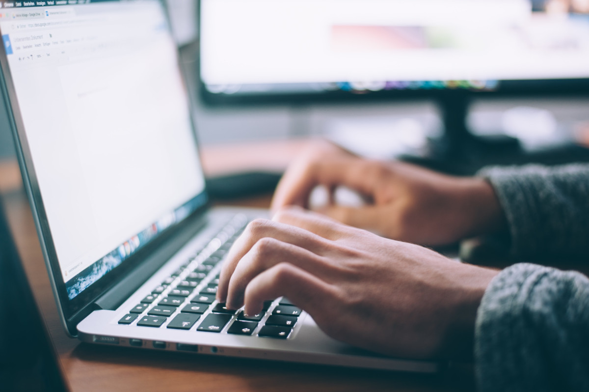 Hands typing on a laptop keyboard, with a blurred computer monitor in the background.