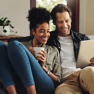 A couple sits on a couch, smiling while looking at a tablet. The woman holds a mug, and a potted plant is in the background.