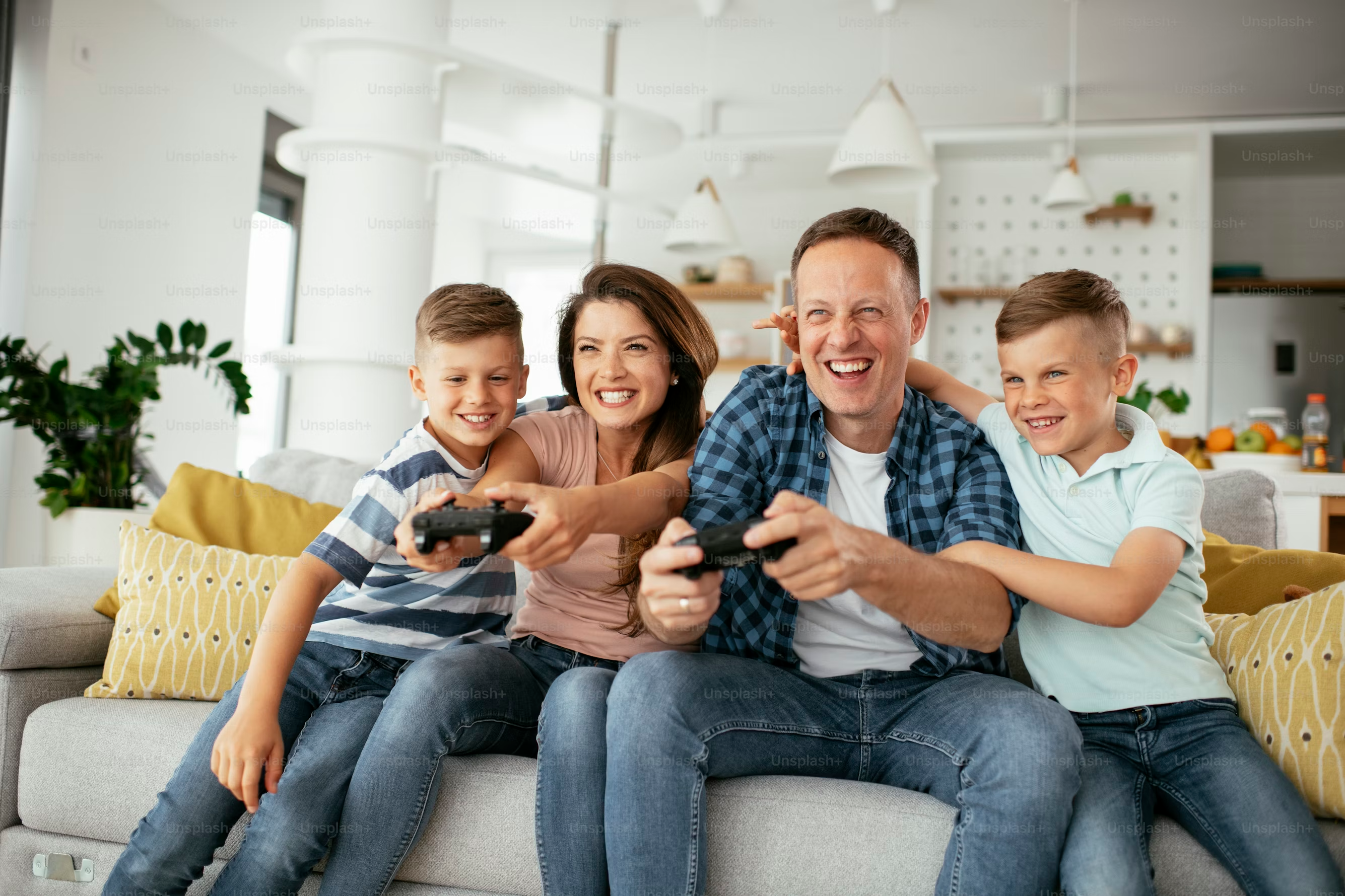 A family of four, two parents and two boys, sits on a couch playing video games, smiling and enjoying their time together in a modern living room.