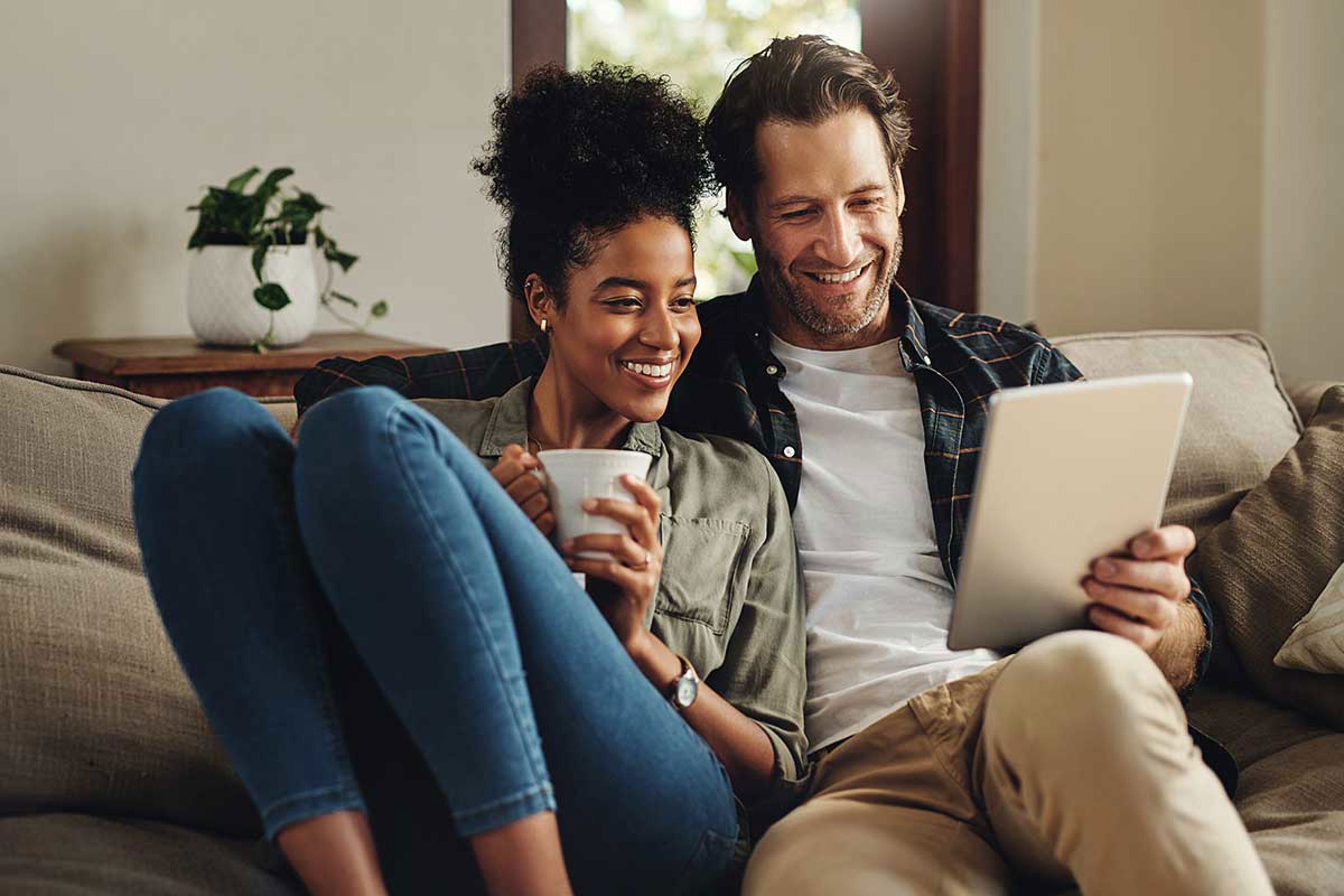 A couple sitting on a couch, smiling while looking at a tablet. The woman is holding a mug, and there