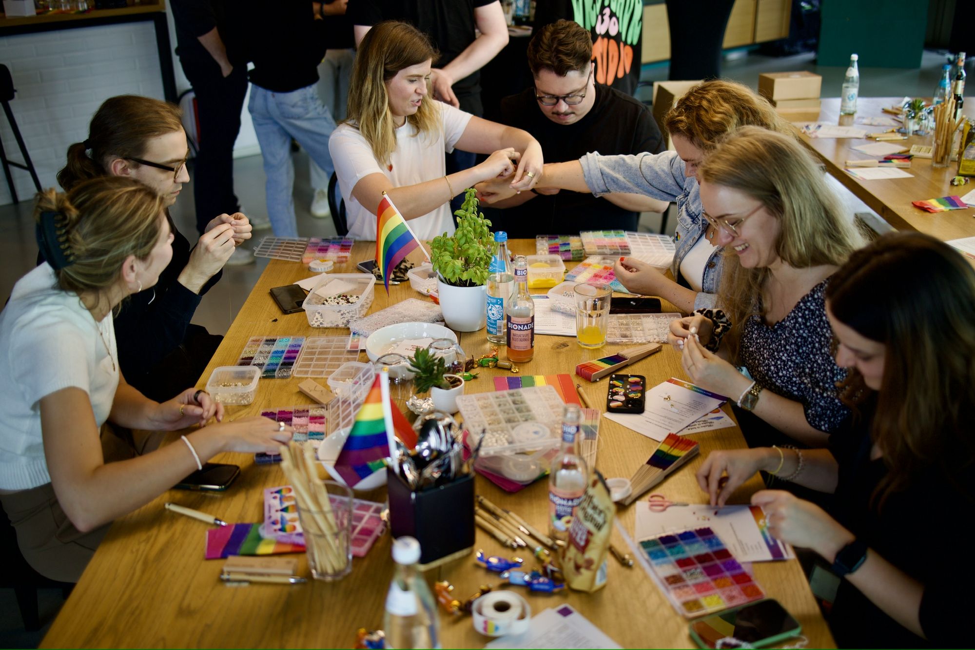 Eine Gruppe von Menschen sitzt an einem Tisch und ist mit Bastelarbeiten beschäftigt. Überall liegen Pride-Flaggen und Kunstbedarf verstreut herum.