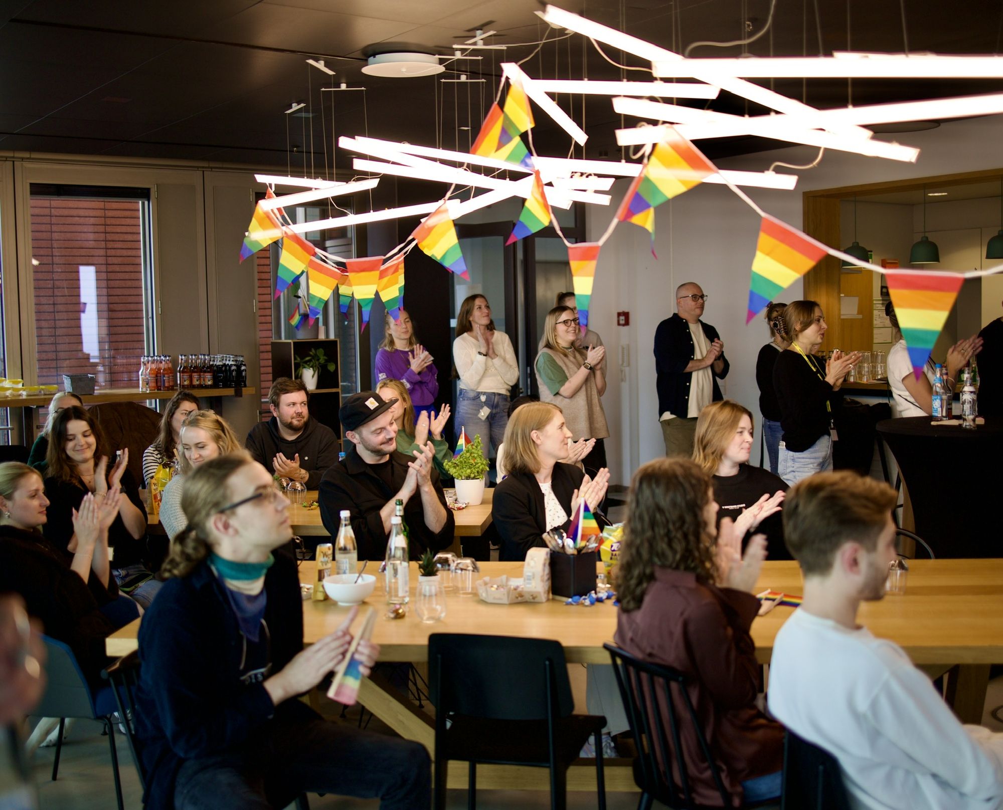 Menschen sitzen in einem mit Regenbogenfahnen geschmückten Raum und verfolgen aufmerksam eine Präsentation, einige klatschen.