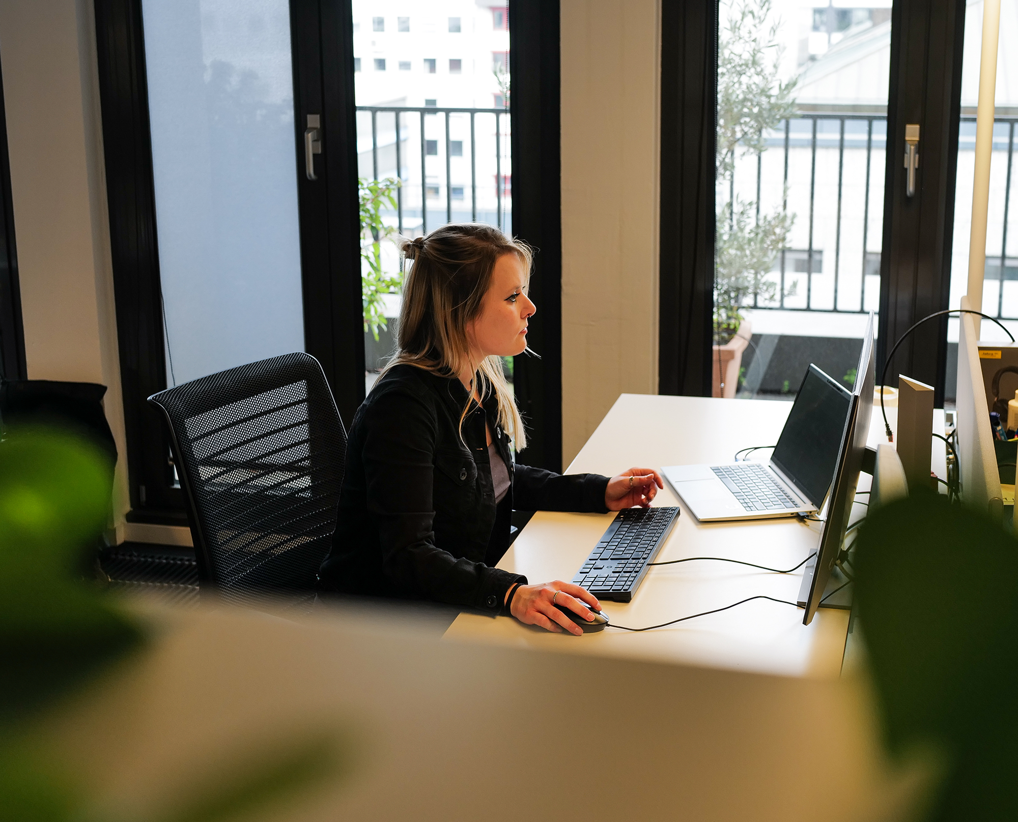 Julia arbeitet an einem Schreibtisch mit Computer und Laptop in einem Büro mit großen Fenstern.