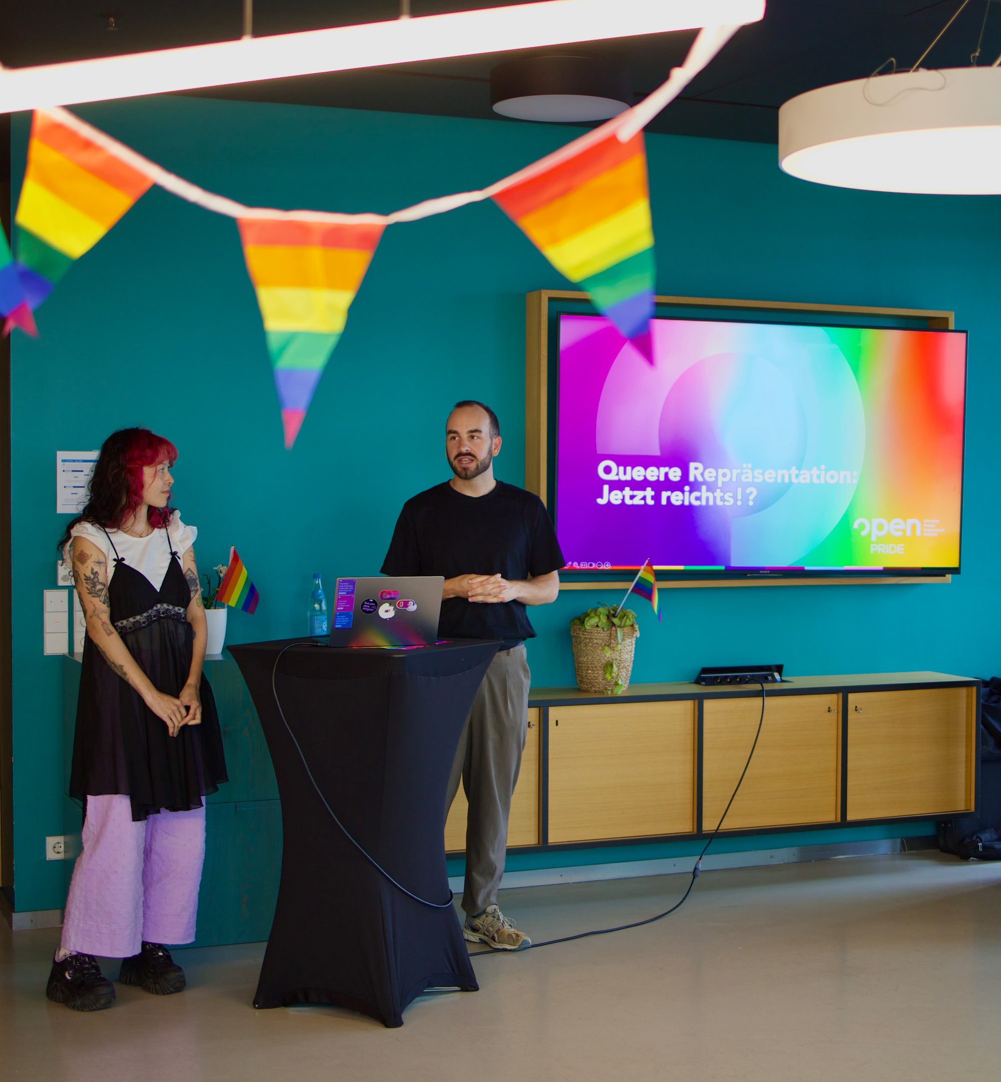 Zwei Menschen stehen neben einem Podium in einem Raum mit Regenbogenfahnen.