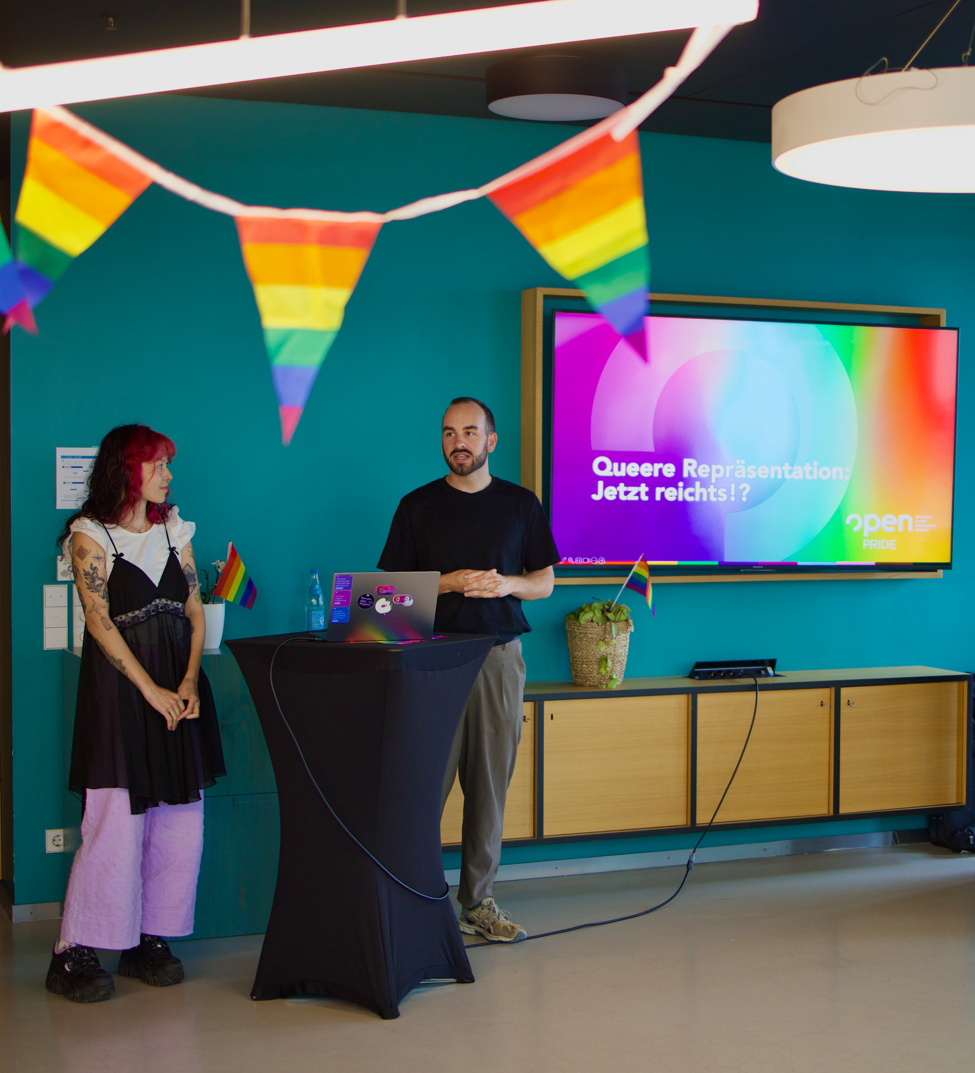 Zwei Menschen stehen neben einem Podium in einem Raum mit Regenbogenfahnen.