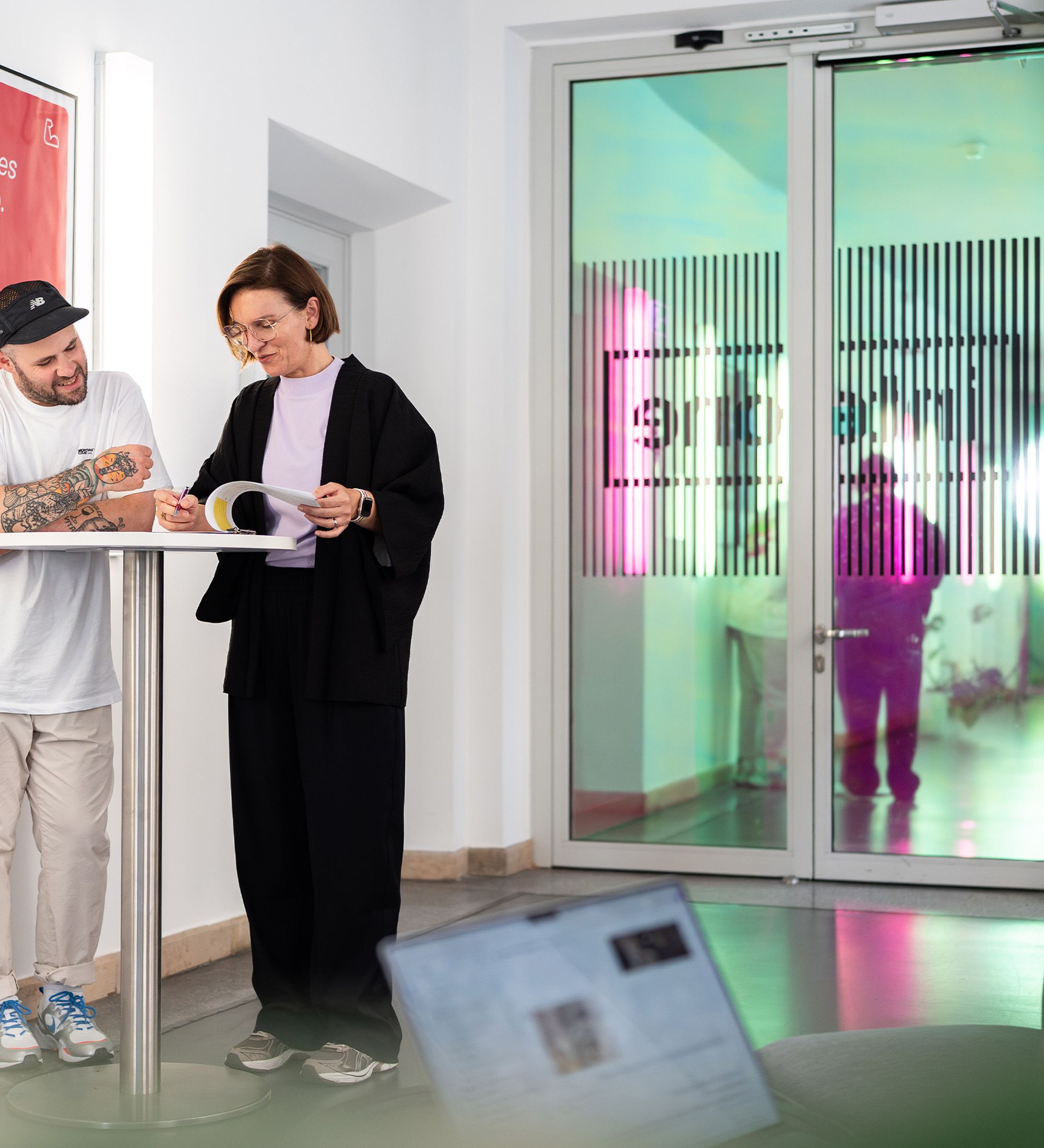 Im Hintergrund sind zwei interone Mitarbeitende im Flur des Münchner Office zu sehen, während sie in einem Katalog blättern. Eine weitere Mitarbeiterin ist unkenntlich in der rechten Ecke am Laptop zu sehen.