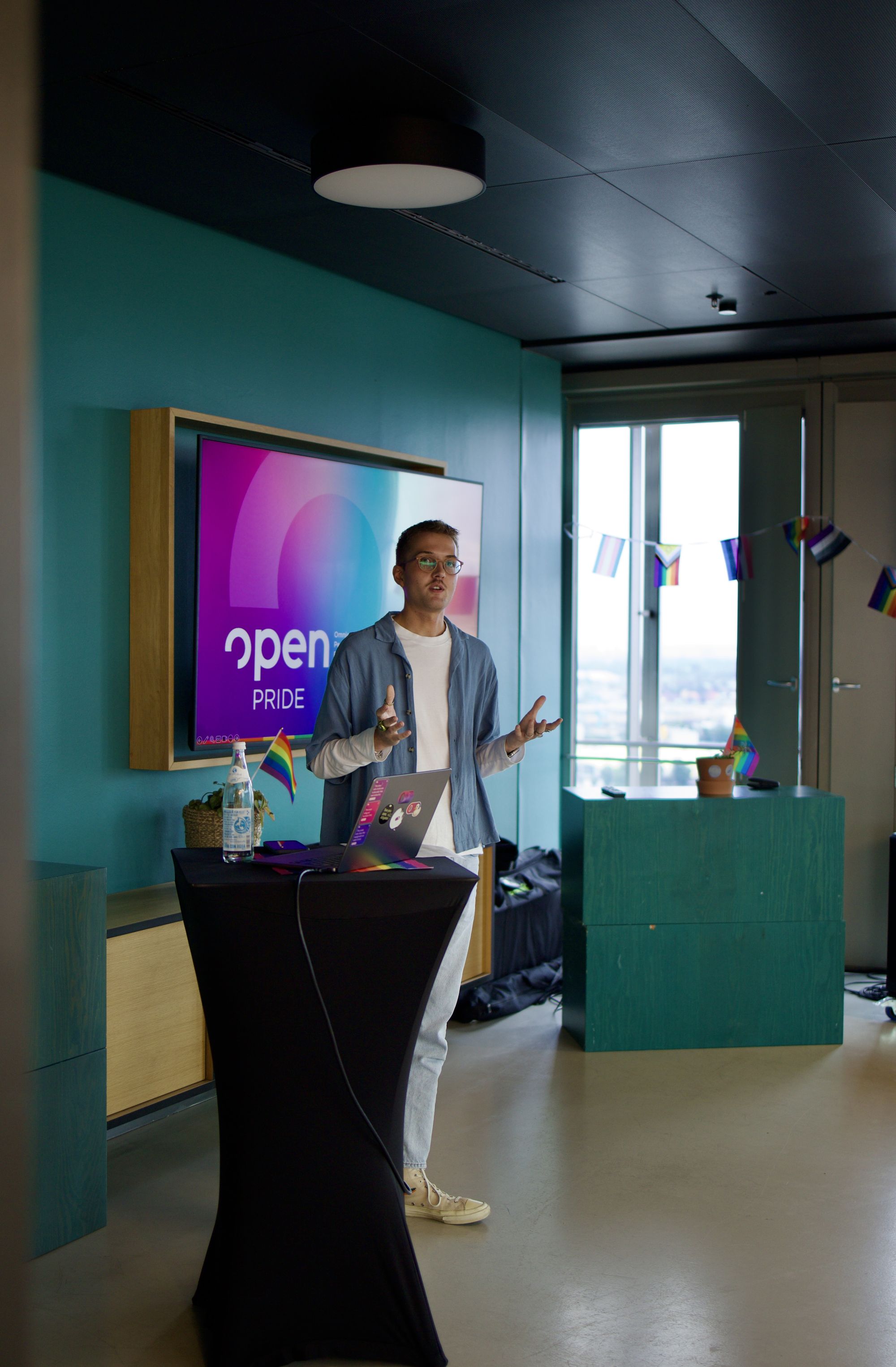 Eine Person spricht mit einem Laptop an einem Podium vor einem Bildschirm mit der Aufschrift „Open PRIDE“. Regenbogenfahnen schmücken den Raum.