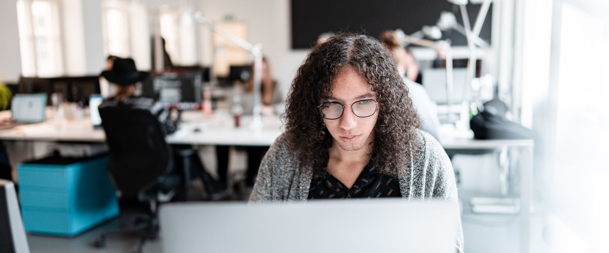 Person mit Laptop arbeitet konzentriert in einem modernen, offenen Büro mit Team im Hintergrund.
