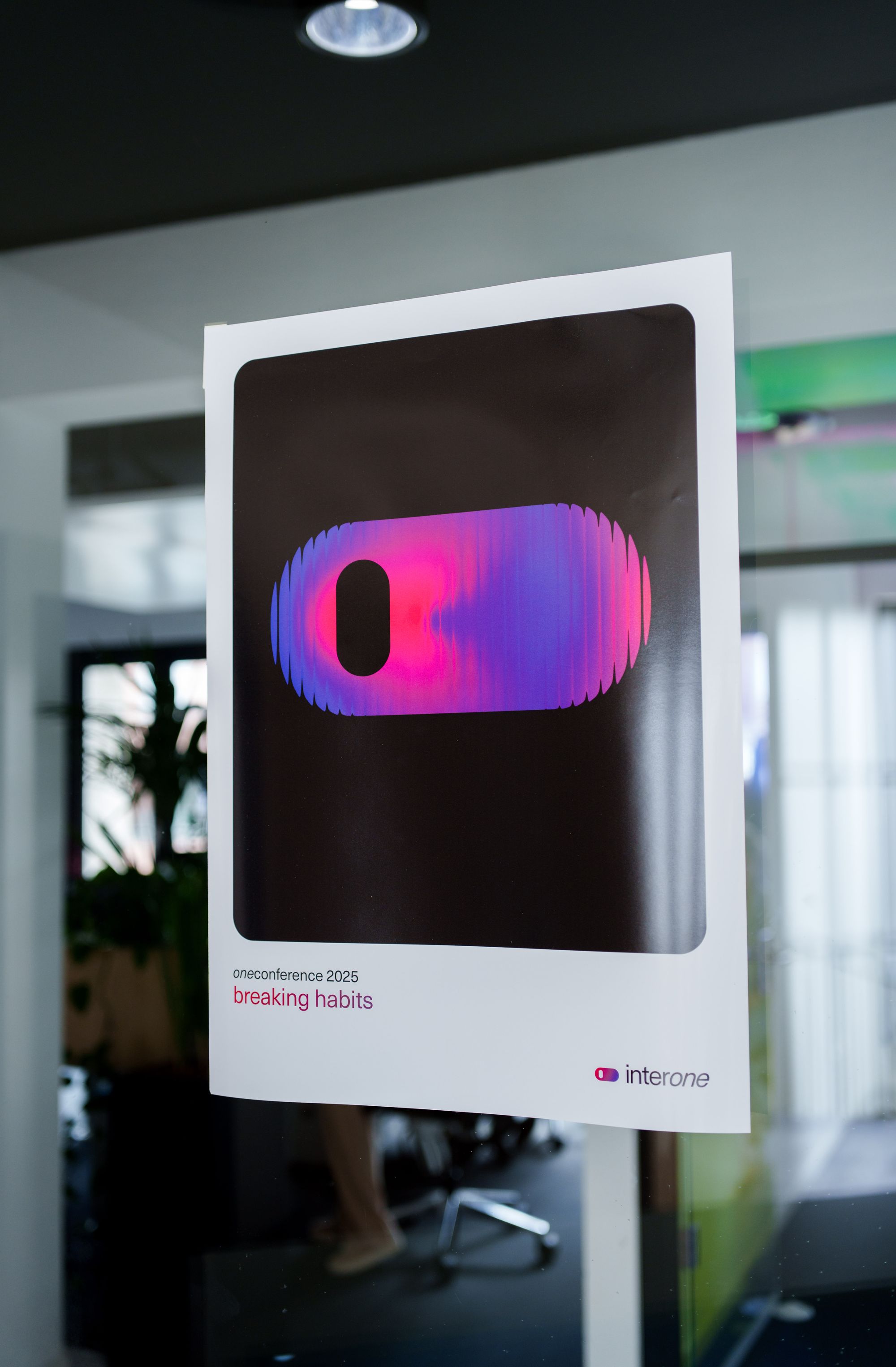 Poster with interone logo in blue, black and pink, labeled "oneconference 2025 breaking habits," displayed in a modern office setting.